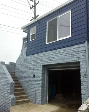 Blue house with garage and stairs. Blue siding, stone facade. Sliding window. Power lines in the background.
