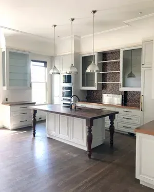 White kitchen with island, cabinets, and three hanging lights. Dark wood countertops and floor.