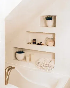 Built-in bathroom shelves with plants, candles, and bath products. White walls and fixtures, gold faucet.