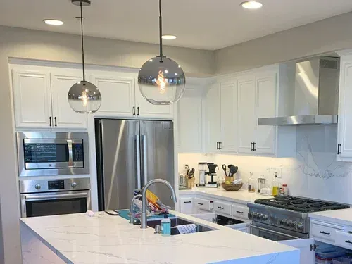 Modern white kitchen with stainless steel appliances, pendant lights, and marble countertops.