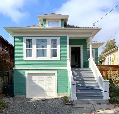 Green house with white trim, garage, stairs, and small dormer under a blue sky.