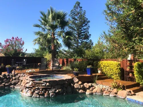 Backyard with a pool, hot tub, and landscaping under a blue sky.