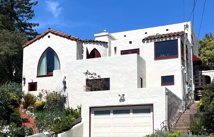 White stucco house with arched window, red trim, and terracotta roof tiles, set on a hillside.