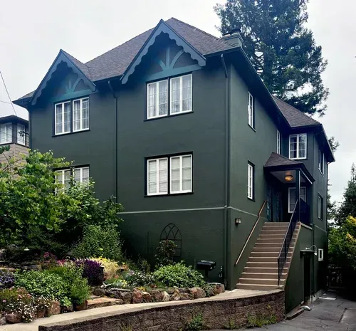 Green two-story house with dark roof and white window trim. Steps lead to a covered entrance. Lush landscaping.