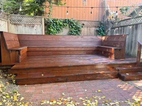 Wooden deck with built-in seating in a backyard. Brown wood, brick patio, ivy on fence.