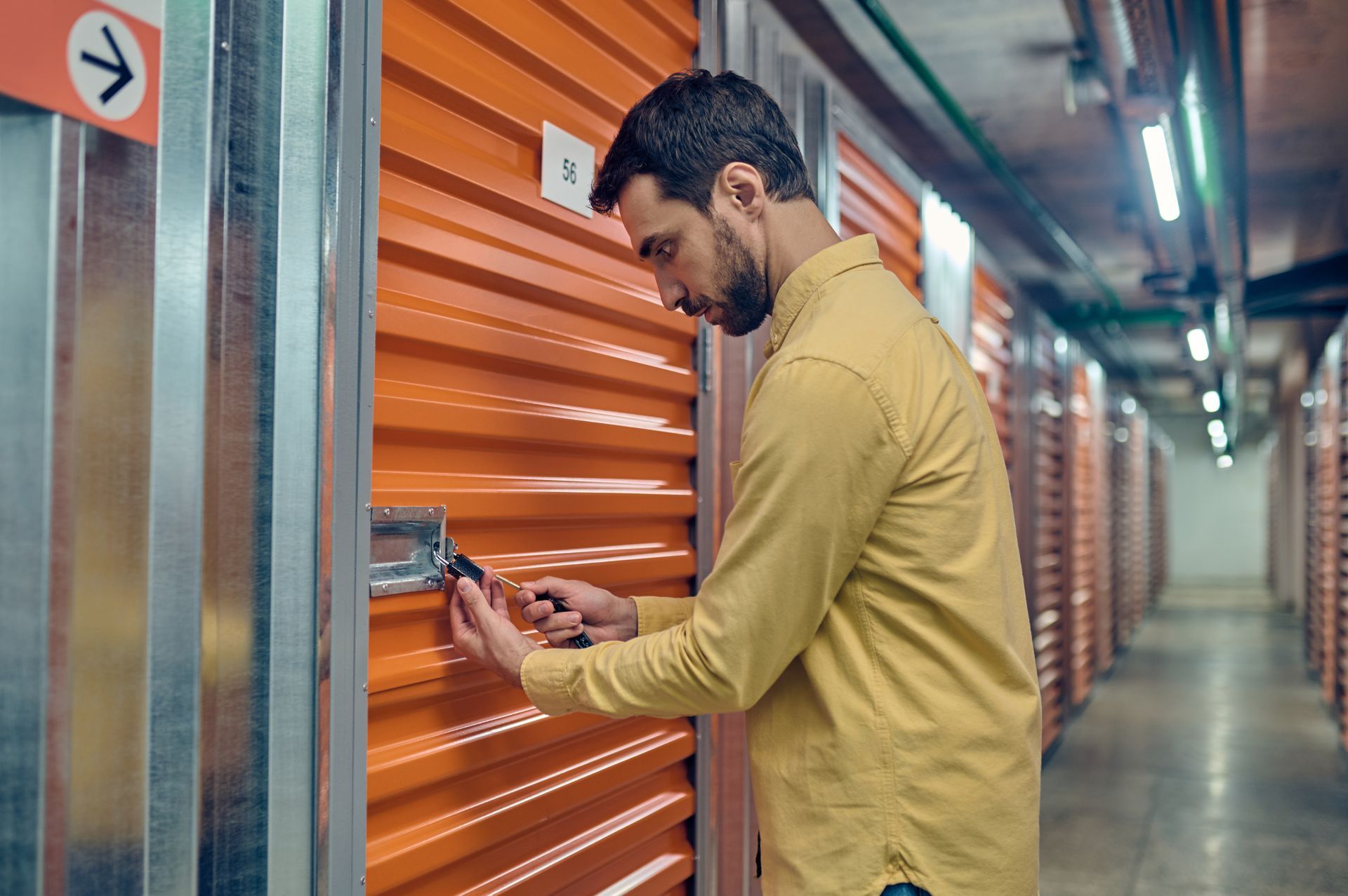 A man unlocking an orange self-storage unit door in an indoor facility hallway.