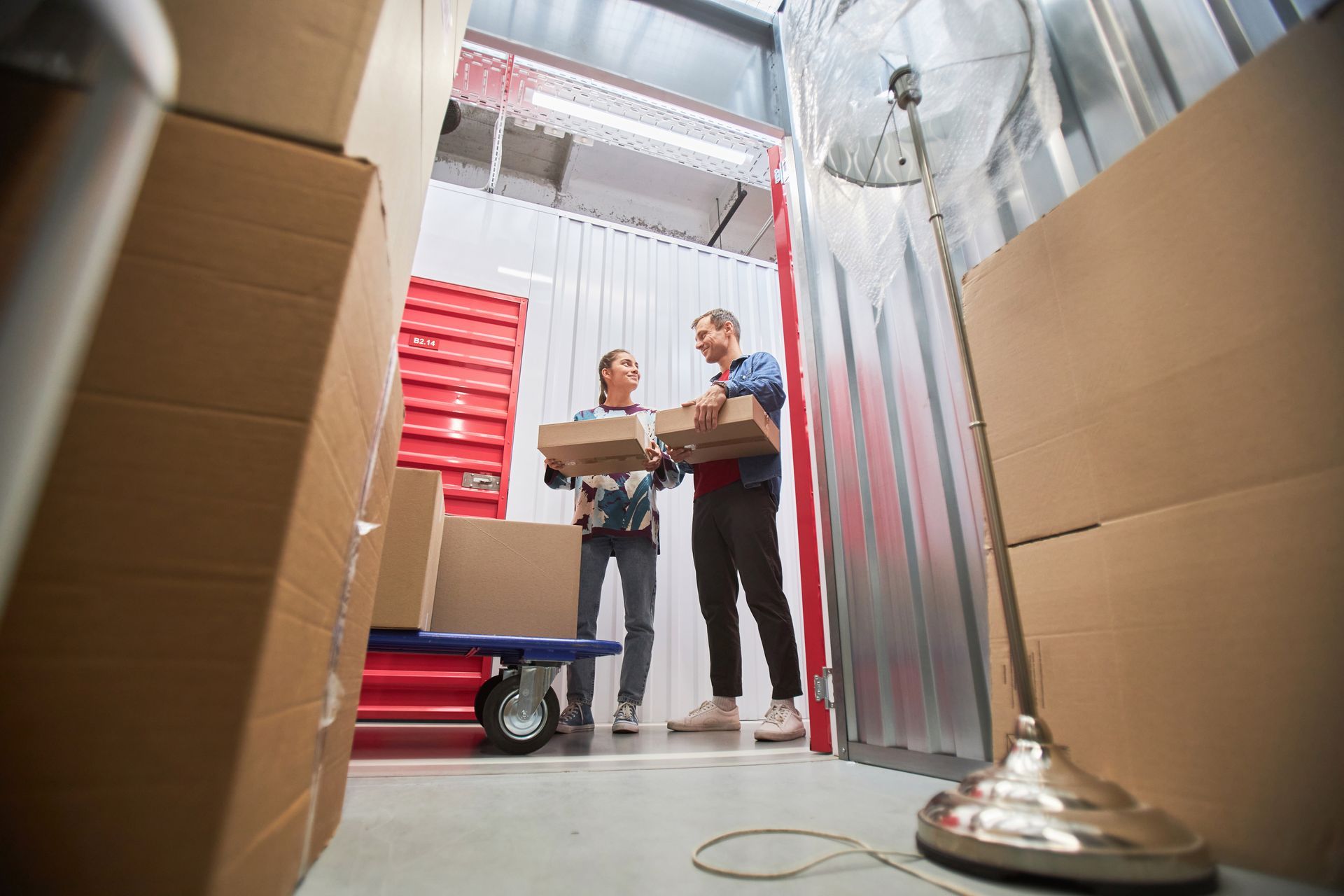 Two people carrying cardboard boxes inside a storage unit with stacked boxes.