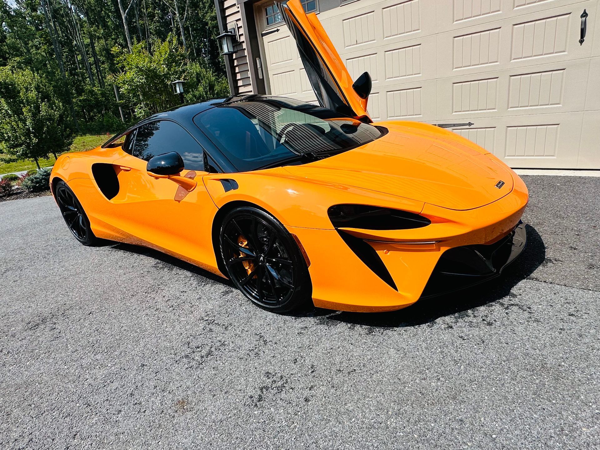 Orange McLaren sports car with black accents parked on gravel driveway, door open.