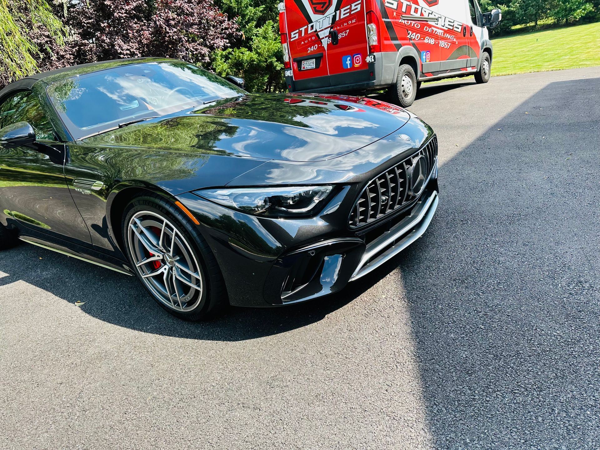 Black Mercedes-Benz convertible parked on asphalt driveway, with a red van in background.