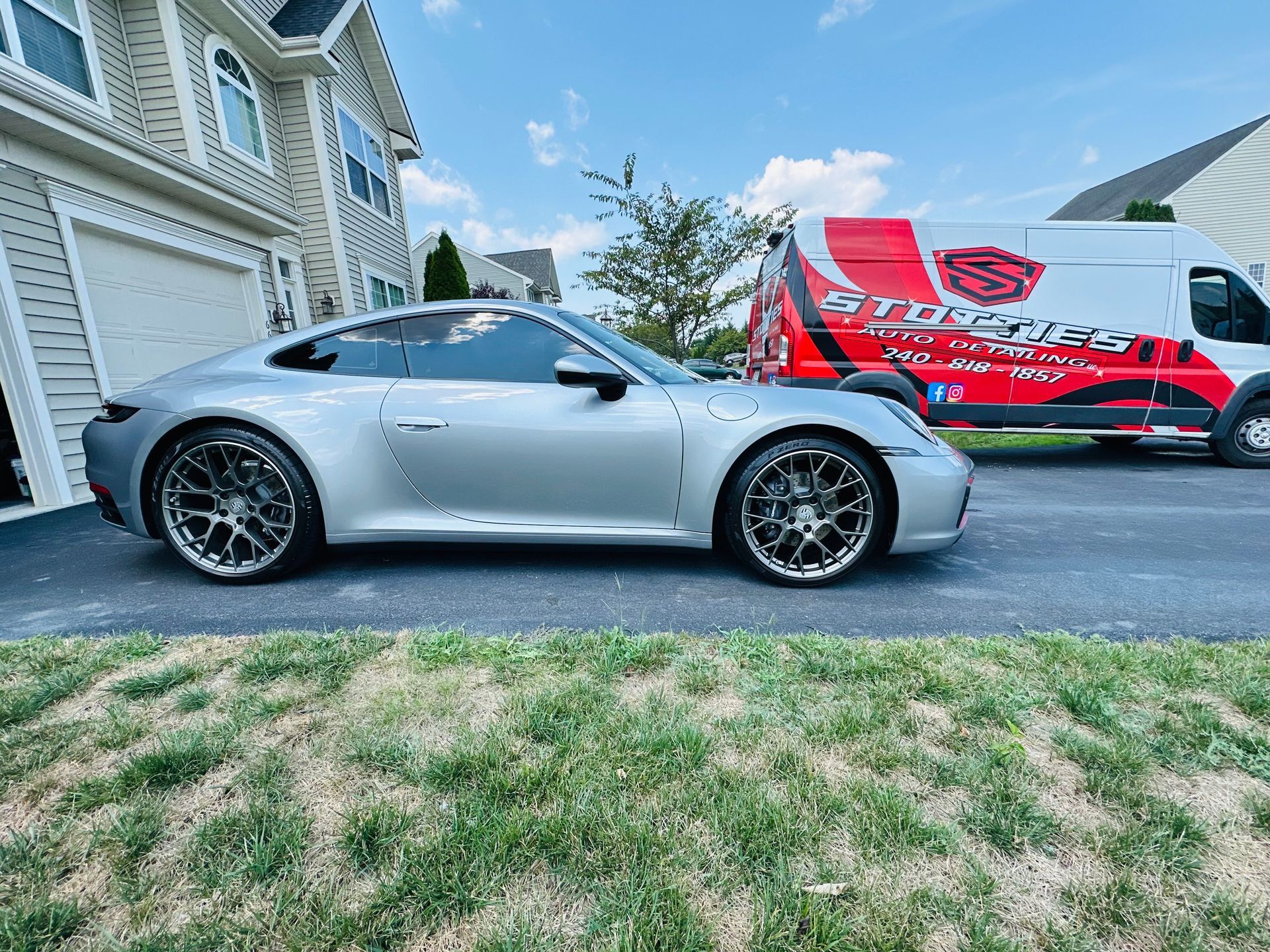 Silver Porsche parked in front of a house next to a branded red and white van.