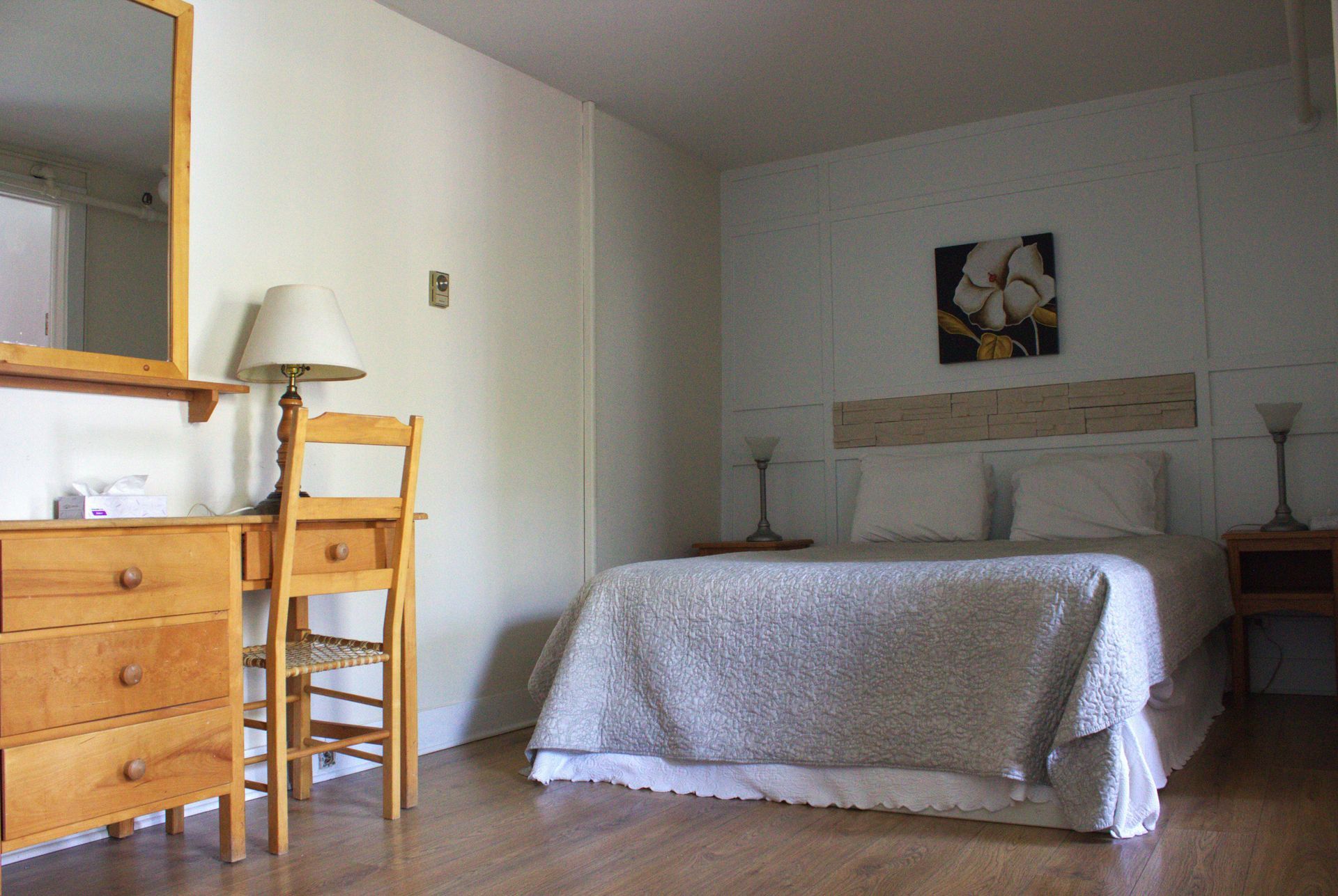 Bedroom with white bedspread, wooden dresser, chair, and lamp in a simple, bright room