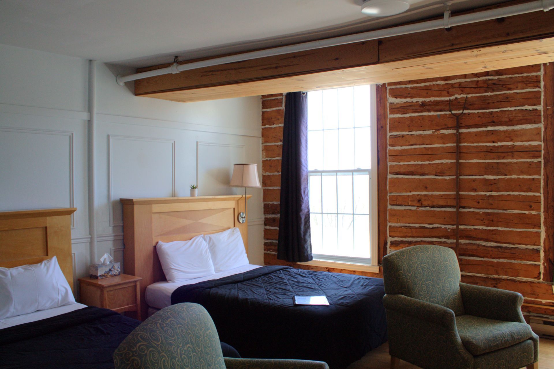 Hotel room with two beds, green chairs, exposed wooden beam, and window with curtains