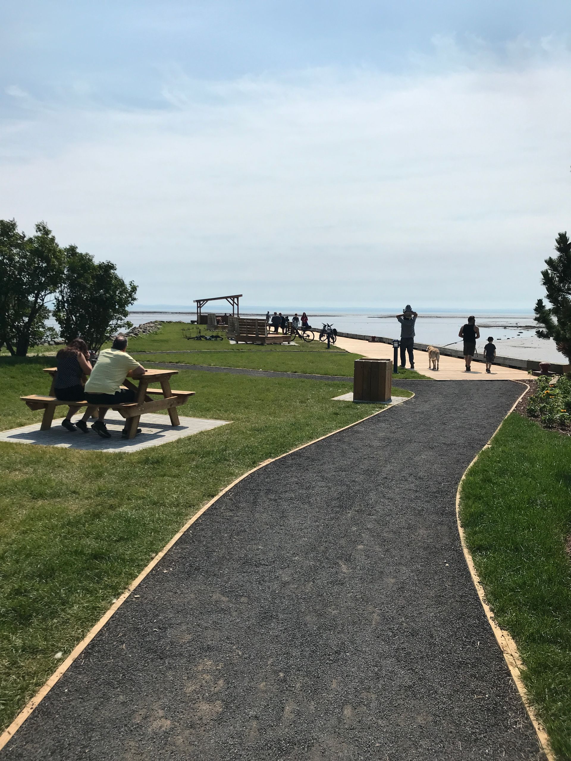 Gravel path leading to a seaside lookout with benches, people, and ocean view under a bright sky