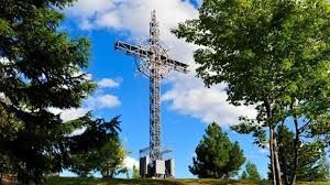 Large metal cross monument in a sunny park, framed by green trees and blue sky.