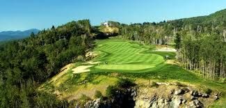Aerial view of a lush green golf course bordered by forest and rocky cliffs under a blue sky
