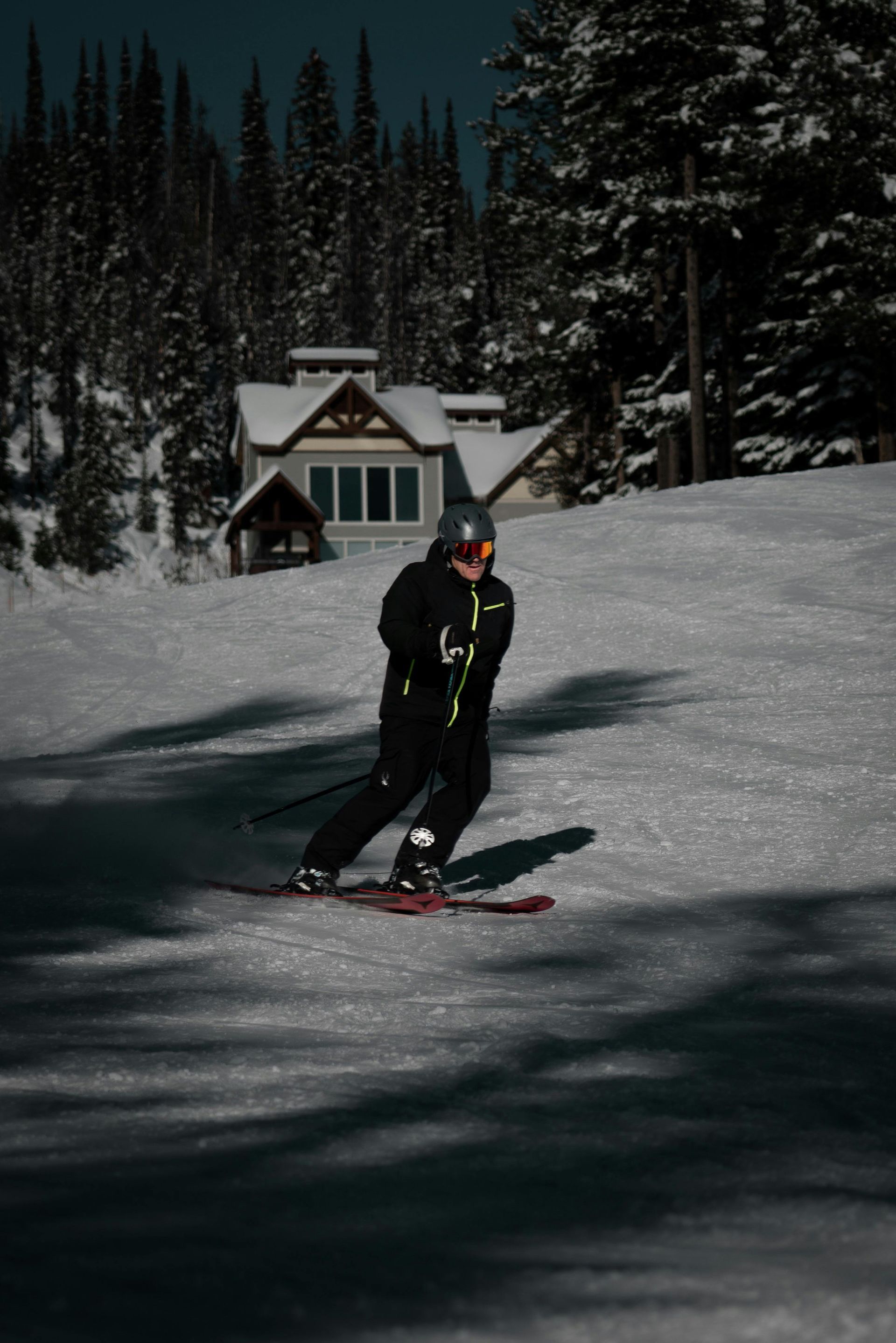 Skier in dark gear skiing down a snowy slope with a cabin and pine trees in the background