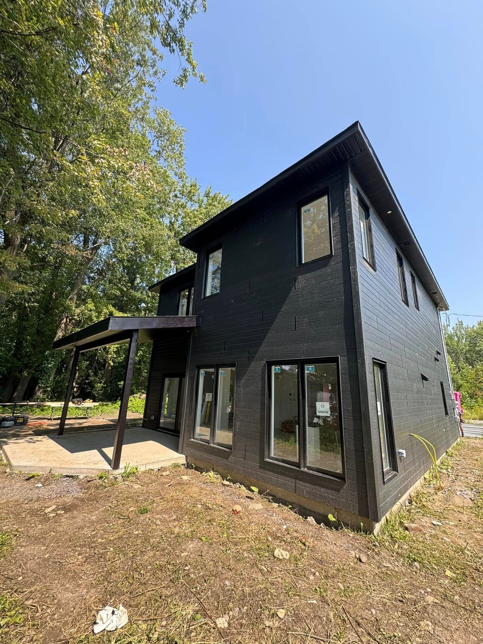 Maison moderne noire avec de grandes fenêtres et un porche couvert, située dans une cour sous un ciel bleu vif.