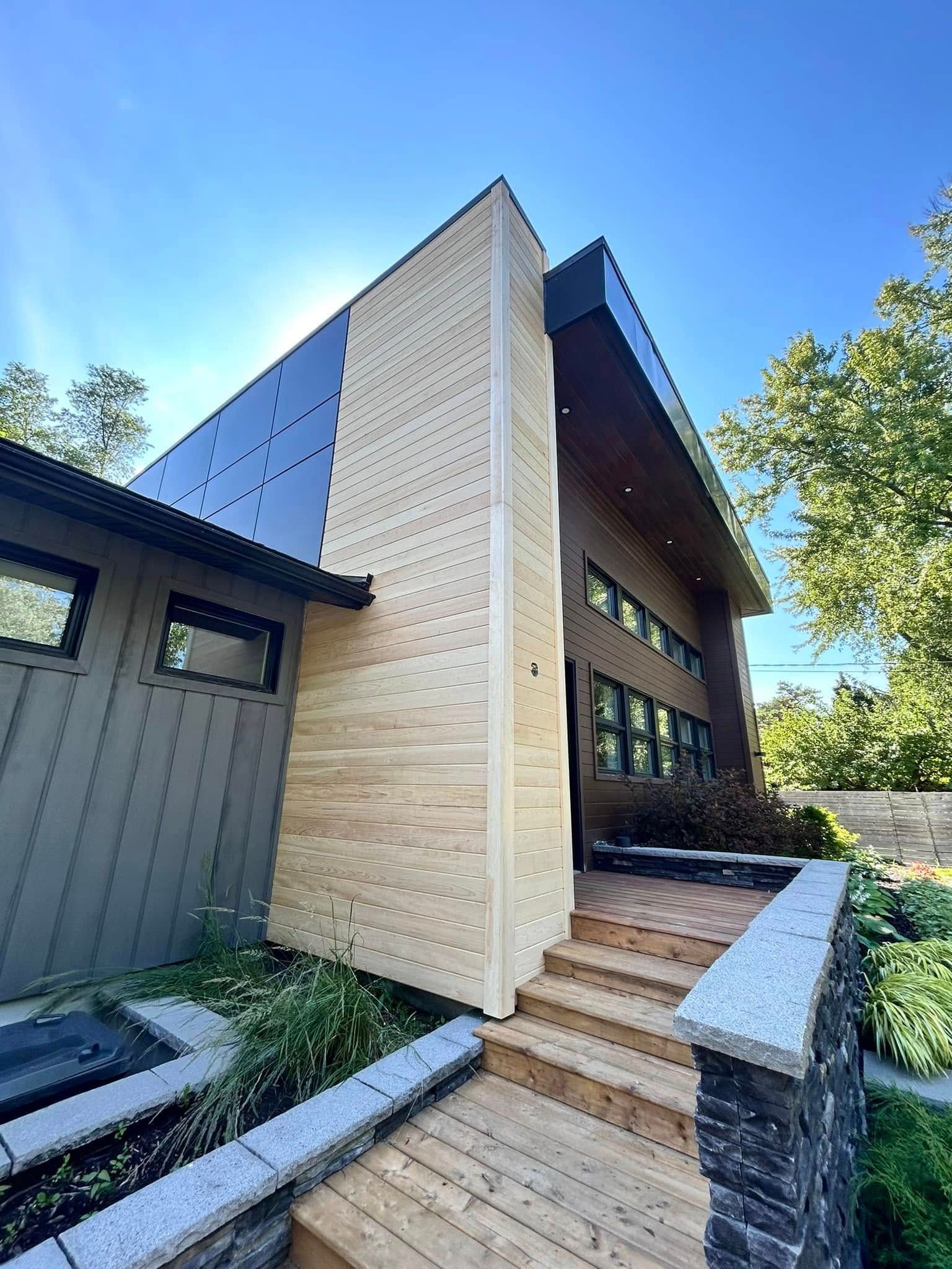 Extérieur d'une maison moderne avec revêtement en bois et foncé, passerelle en bois et ciel ensoleillé.