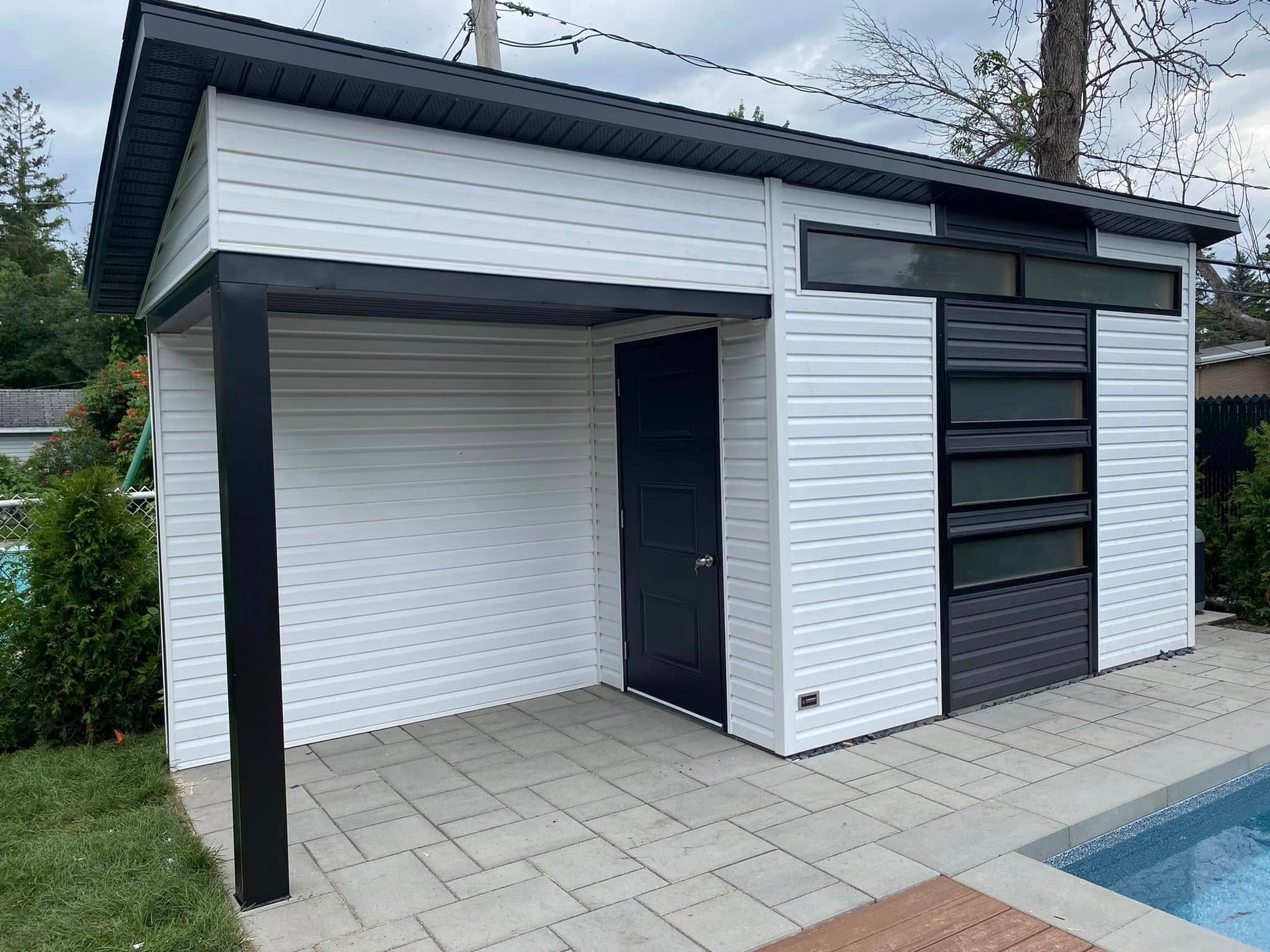 Cabanon blanc aux accents noirs, construit près d'une piscine, avec une porte et des fenêtres foncées.