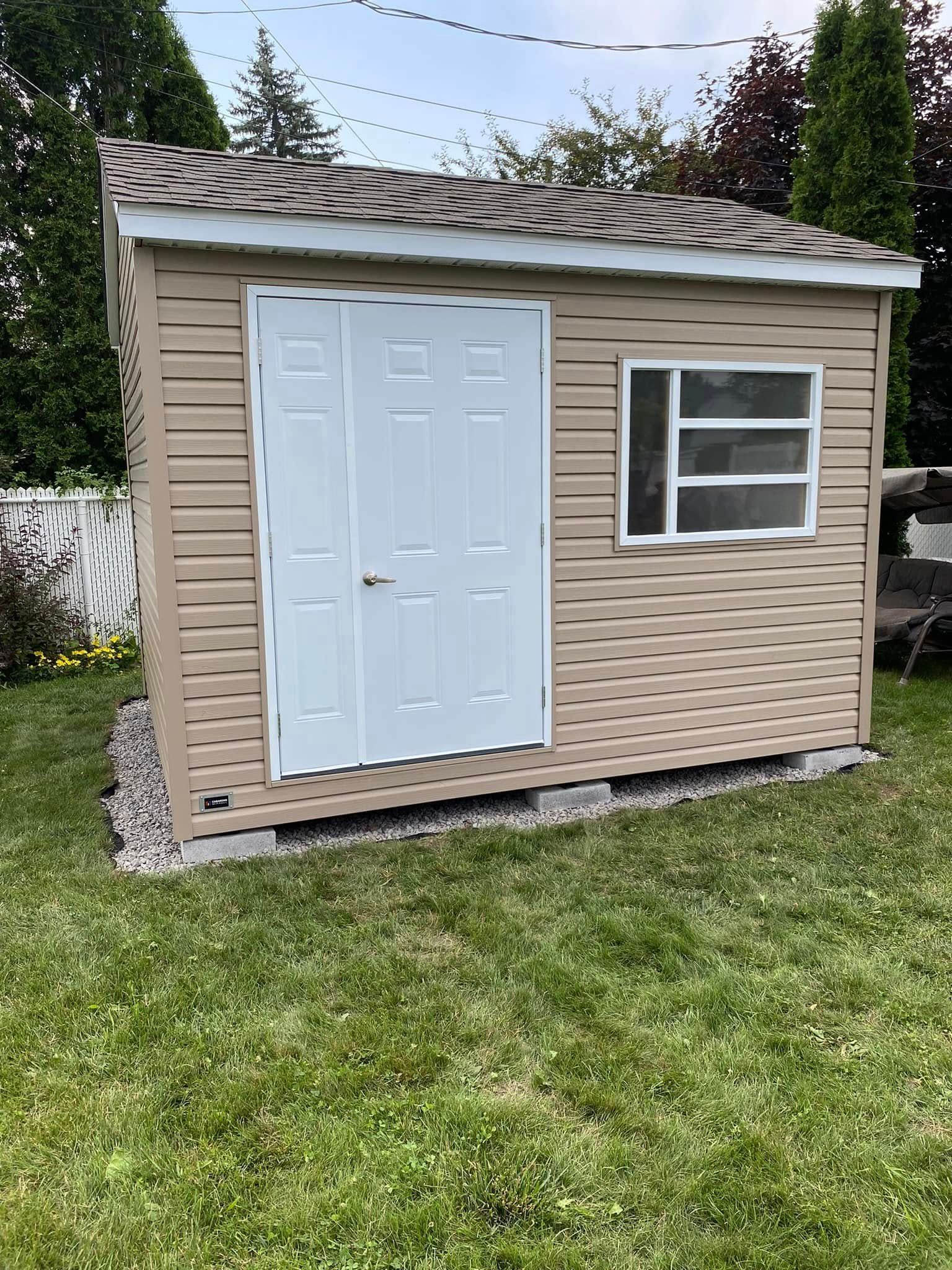 Un petit hangar avec une fenêtre se trouve au milieu d'une cour verdoyante.