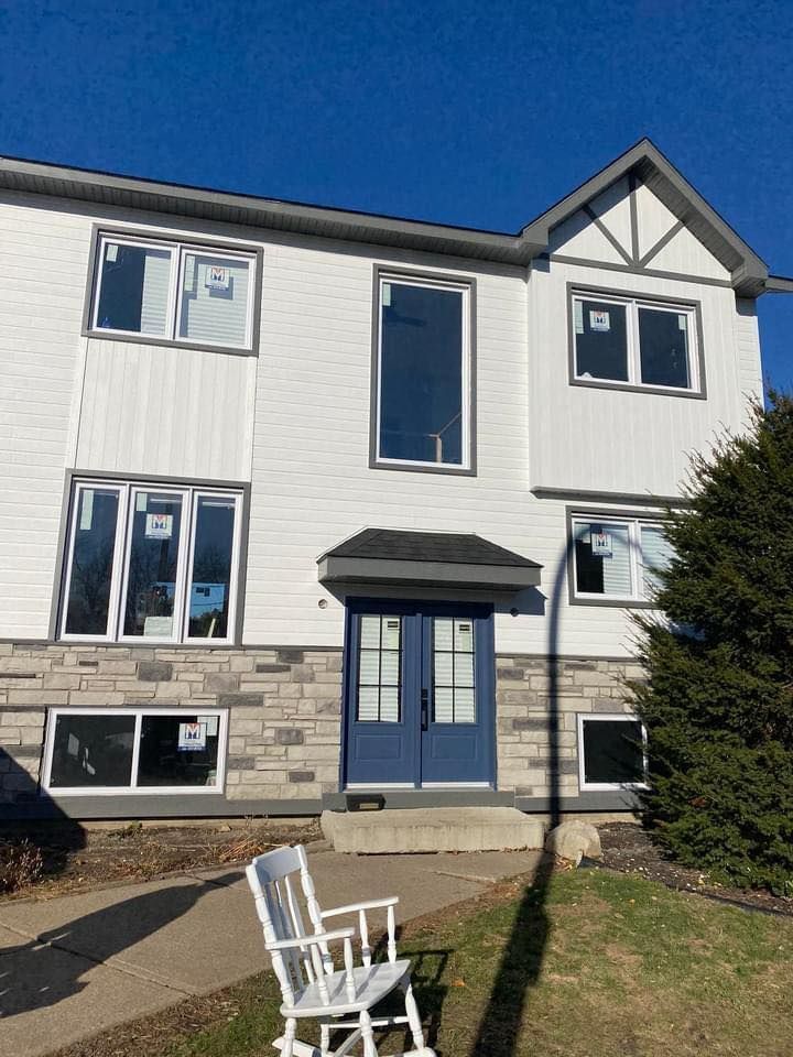 Maison à deux étages avec bardage blanc, porte bleue et façade en pierre. Un rocking-chair blanc trône sur les marches de l'entrée.