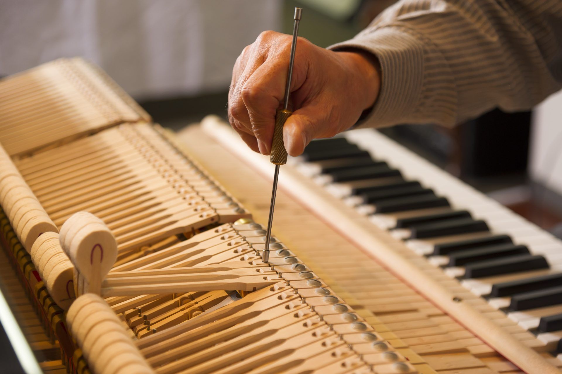 Hand of a person tuning a piano with a tool, focusing on the mechanism, keyboard in background.