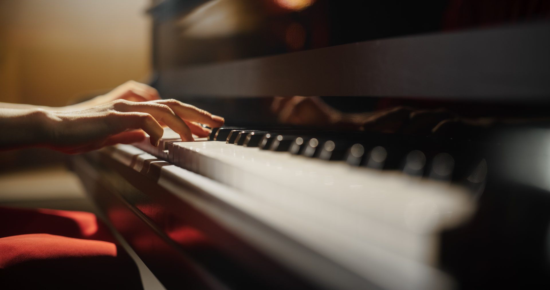Hands playing a piano, close-up view. Black and white keys; red seat. Warm lighting.