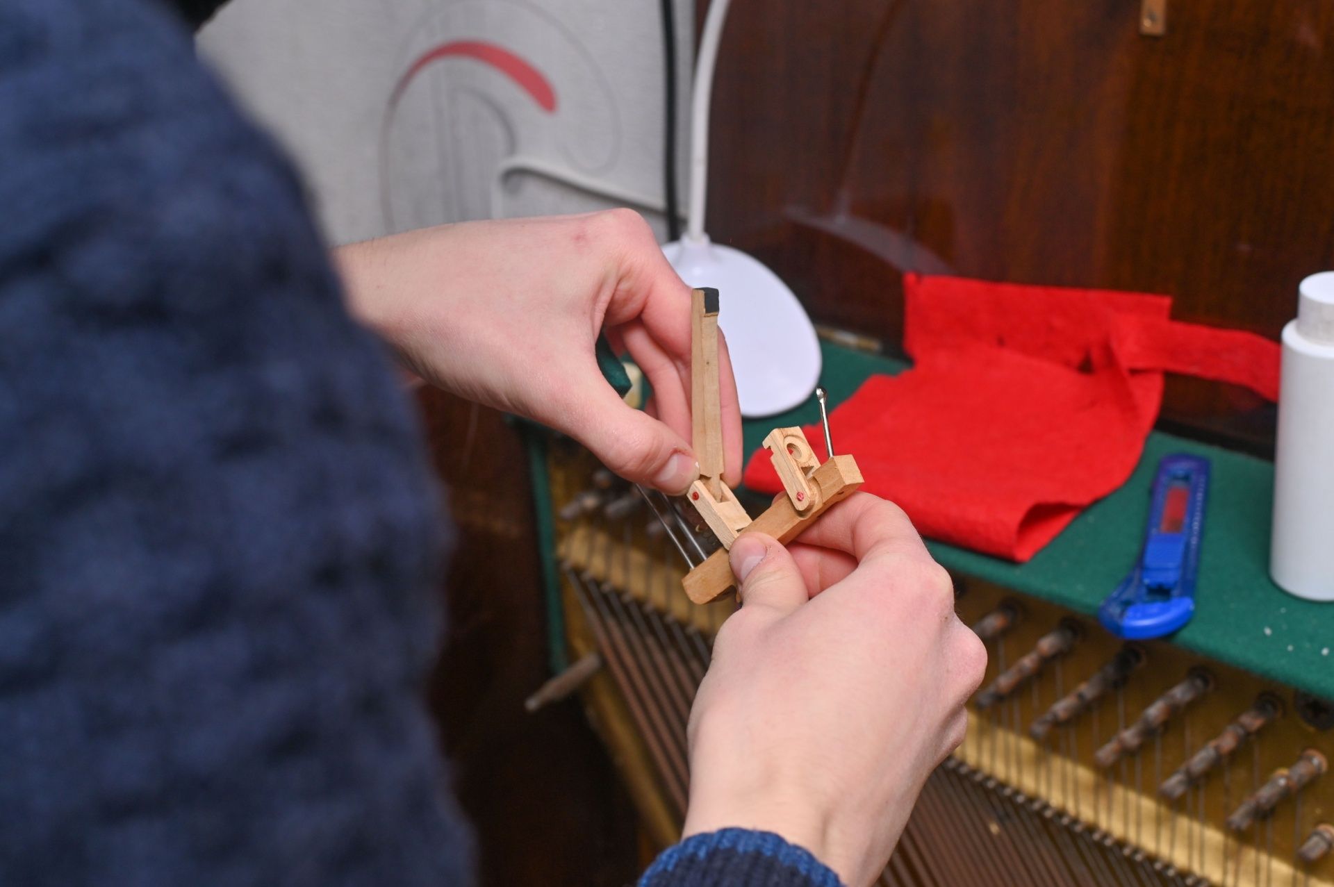 Person repairing piano mechanism, holding wooden parts over piano strings, with tools and a red cloth nearby.