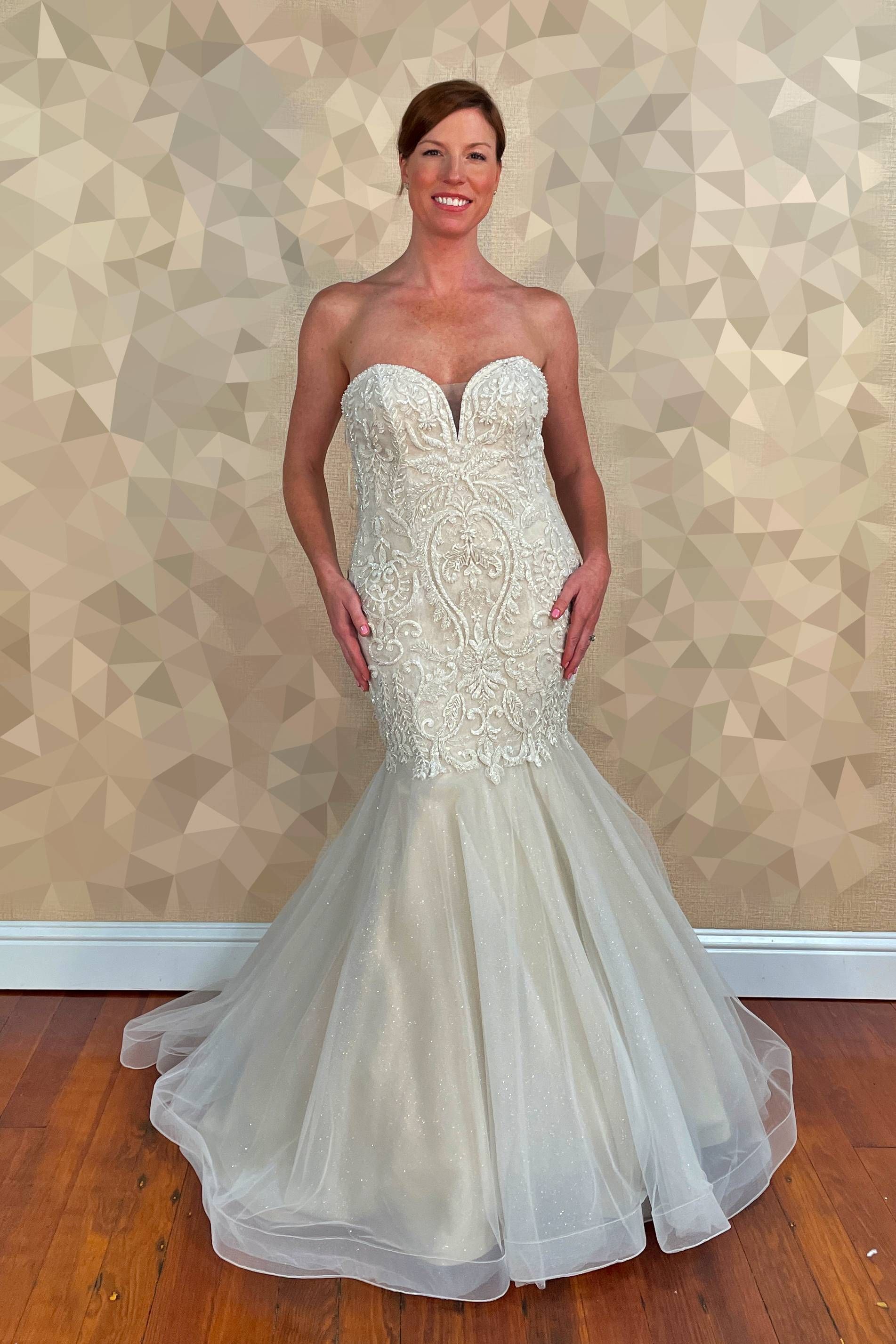 Woman in a strapless beaded mermaid wedding dress stands in front of a patterned wall and wood floor.