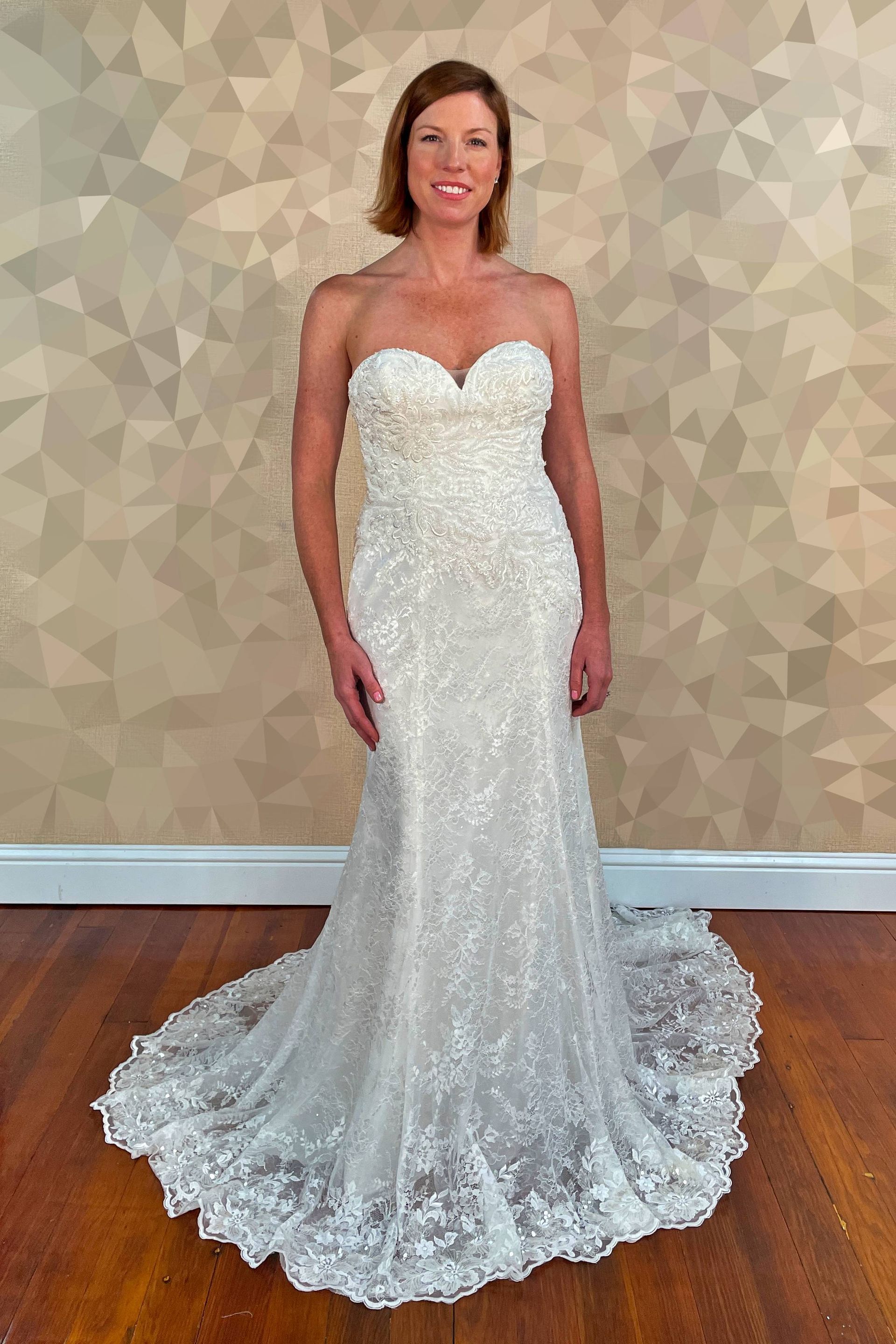 Woman in a white, embellished strapless wedding dress, smiling. Stands in front of textured wall, wooden floor.