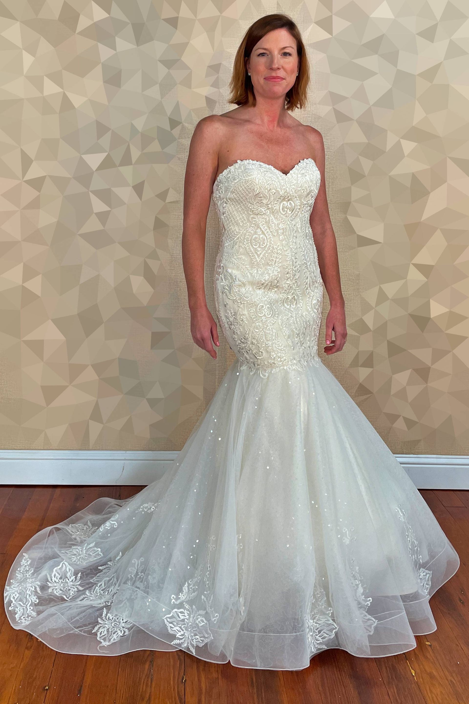 Woman wearing a strapless, embellished white wedding gown with a mermaid silhouette, standing in front of a patterned wall.