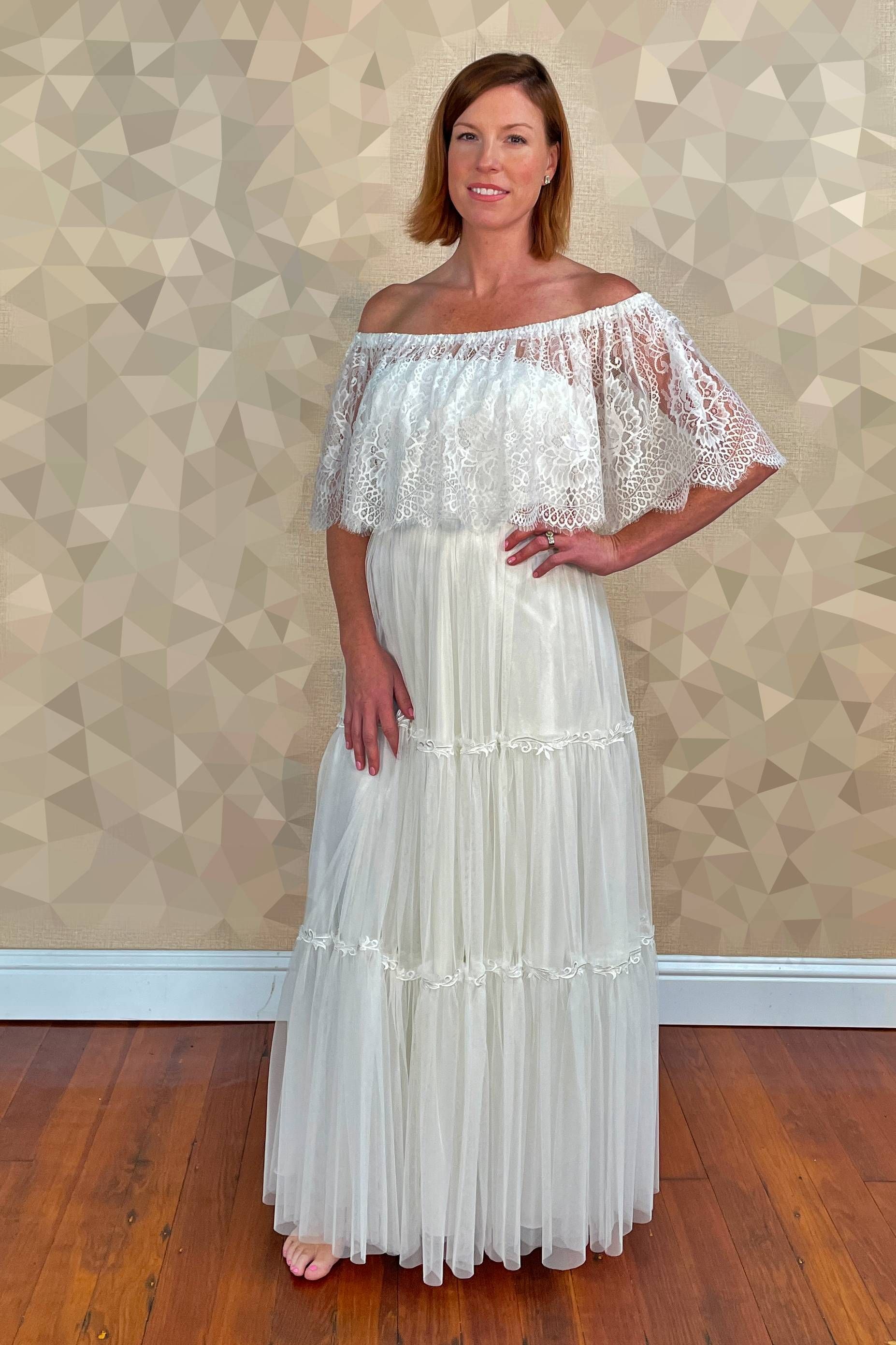 Woman in white off-shoulder lace dress, standing with hand on hip, against a patterned wall.
