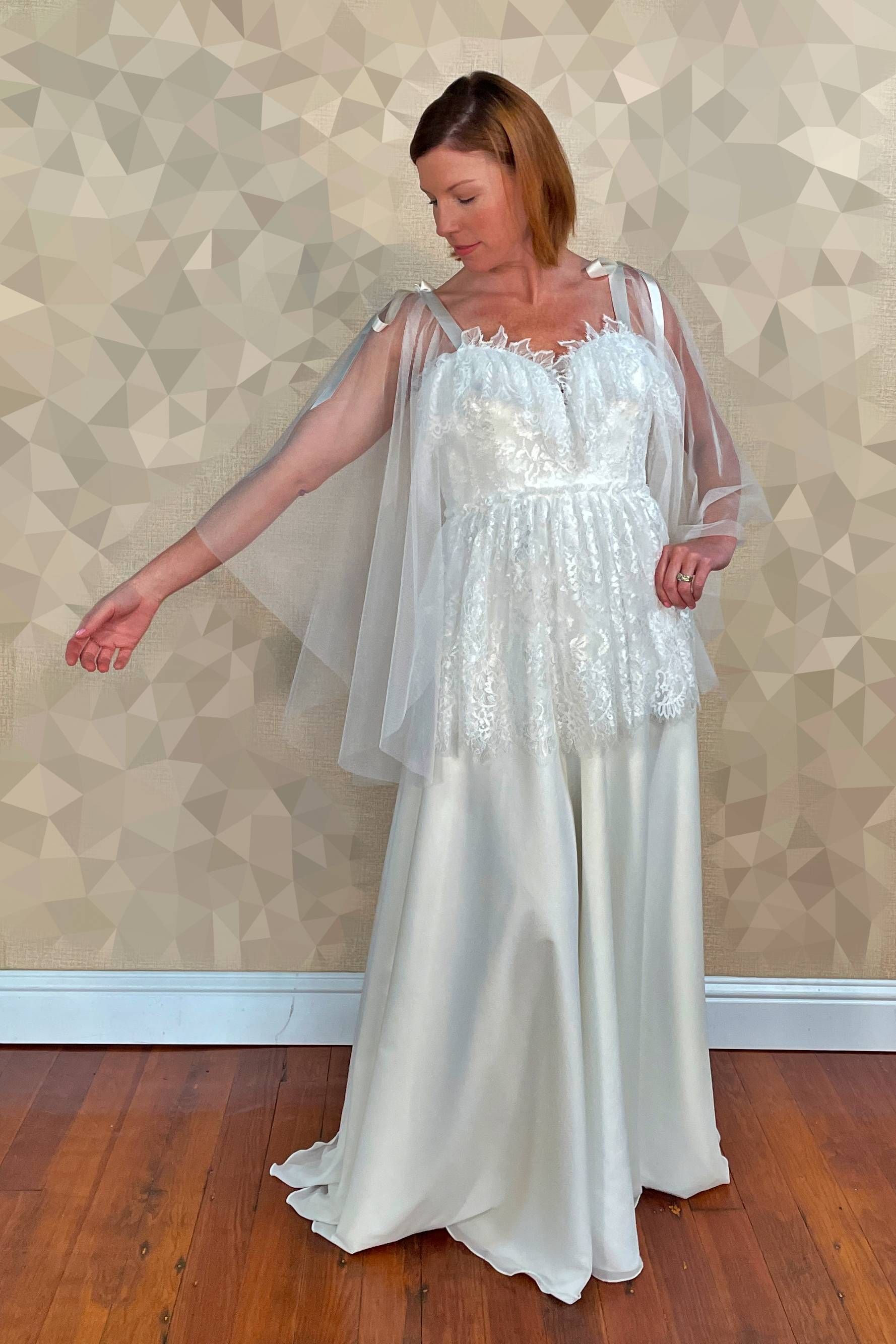 Woman in a white wedding jumpsuit with a sheer cape; stands indoors with one arm extended.