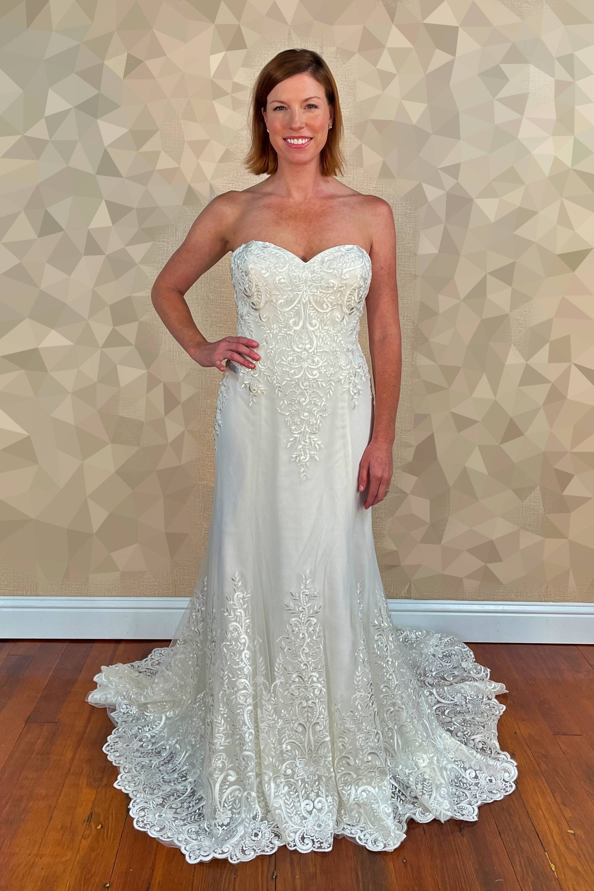 Woman in white strapless wedding gown, posing in a room with patterned walls and hardwood floors.