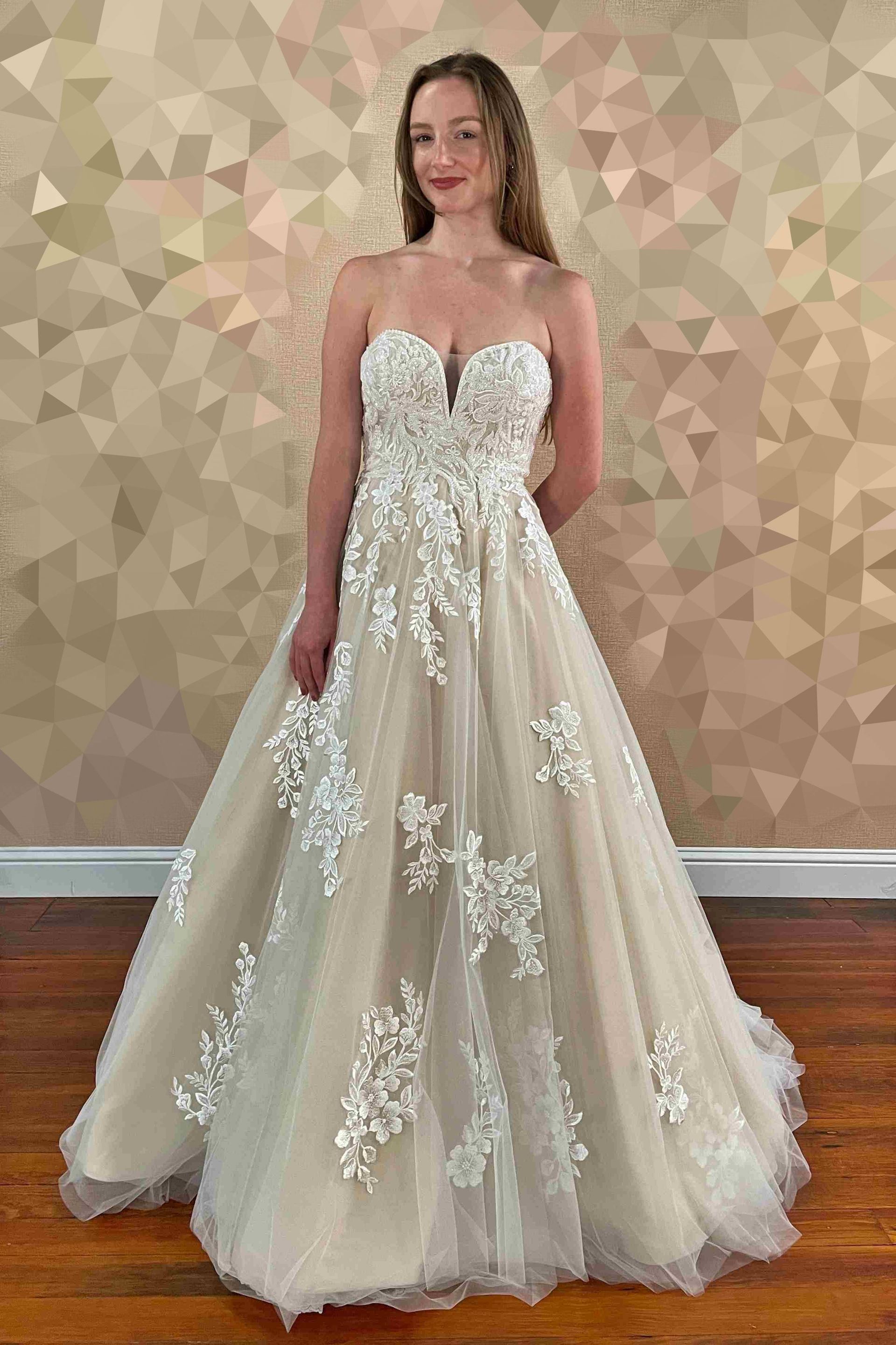 Woman in a strapless ivory and beige wedding gown, with floral embellishments, posing in a room.