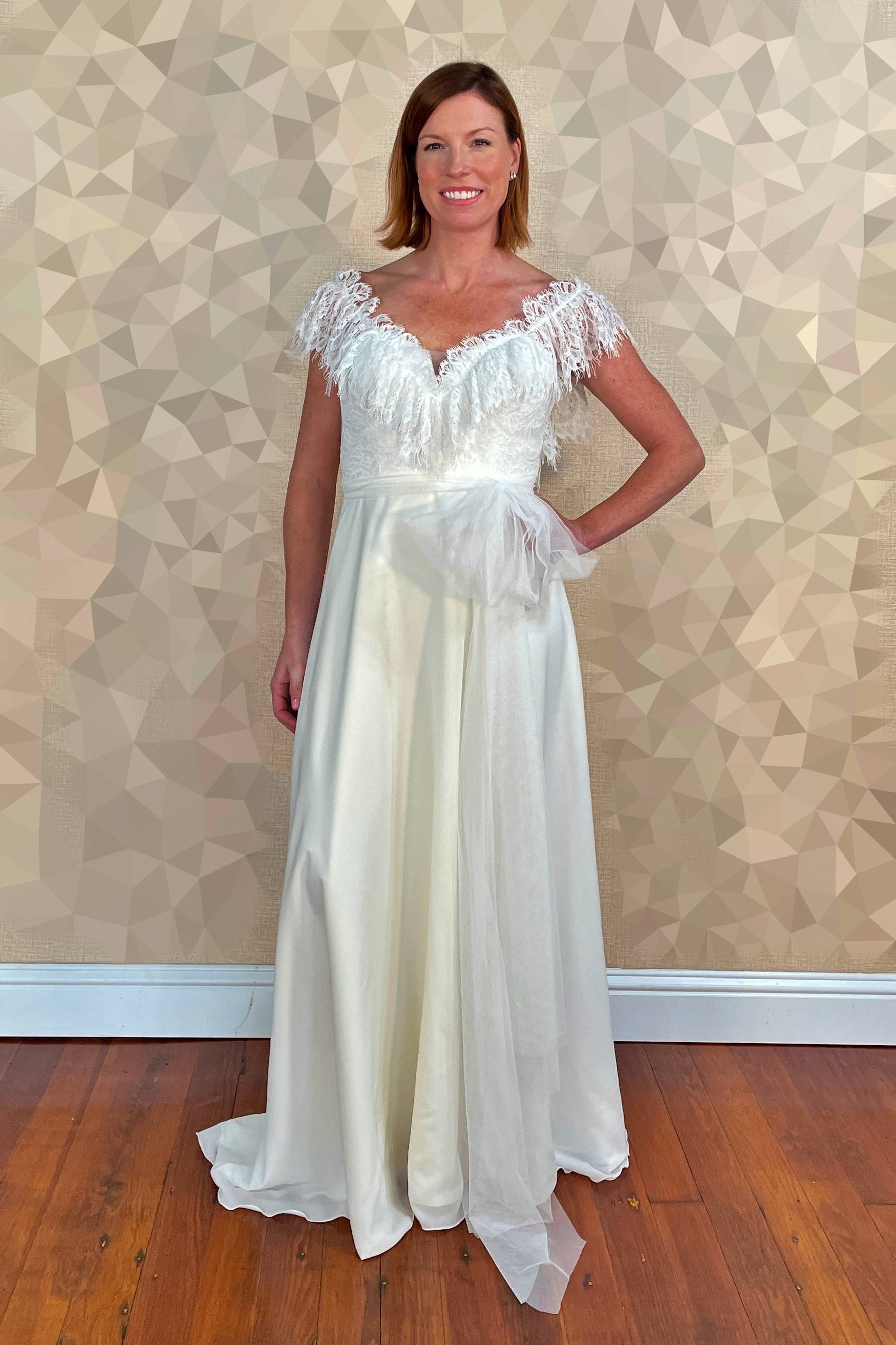 Woman in white dress with lace detail stands, hand on hip, against a gold patterned wall.