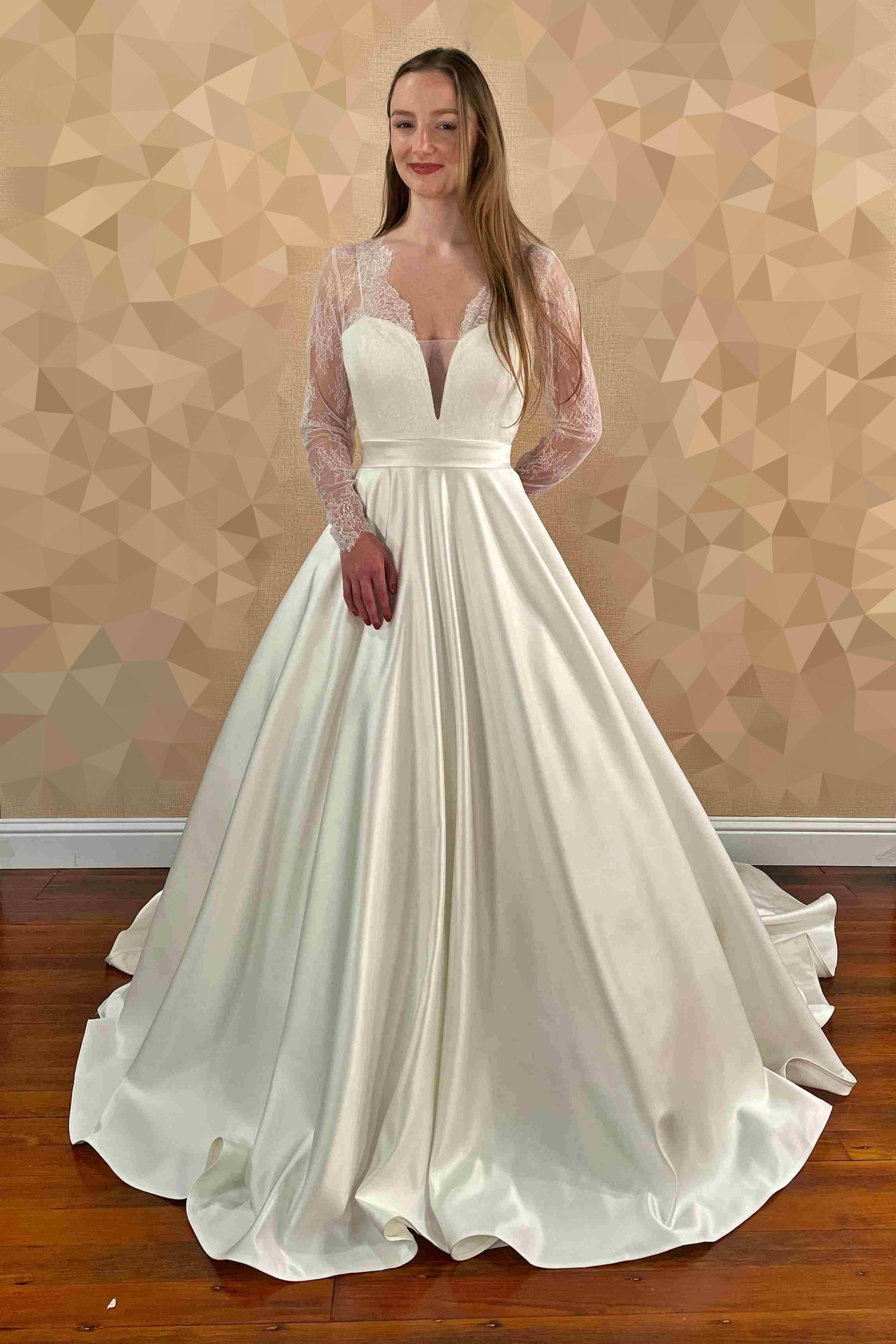 Woman in a white wedding dress with long lace sleeves and a full skirt, standing indoors.