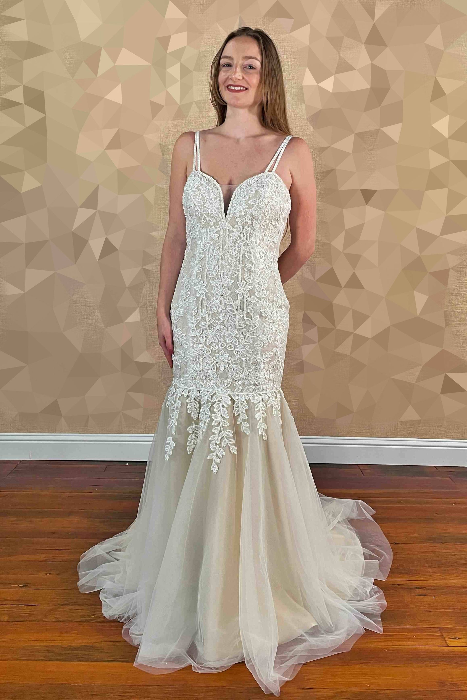 Woman in a white wedding dress with lace bodice, spaghetti straps, and mermaid skirt, smiling indoors.