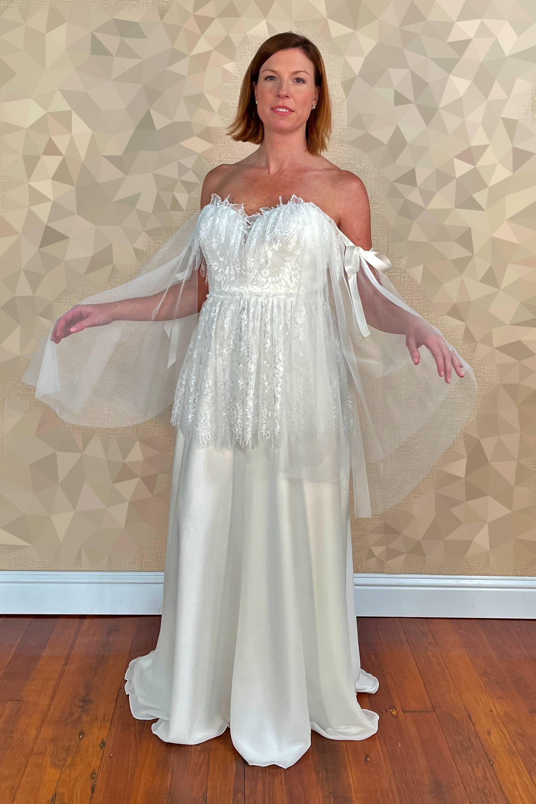 Woman in off-the-shoulder white dress with sheer cape, arms outstretched. Standing in front of textured wall.