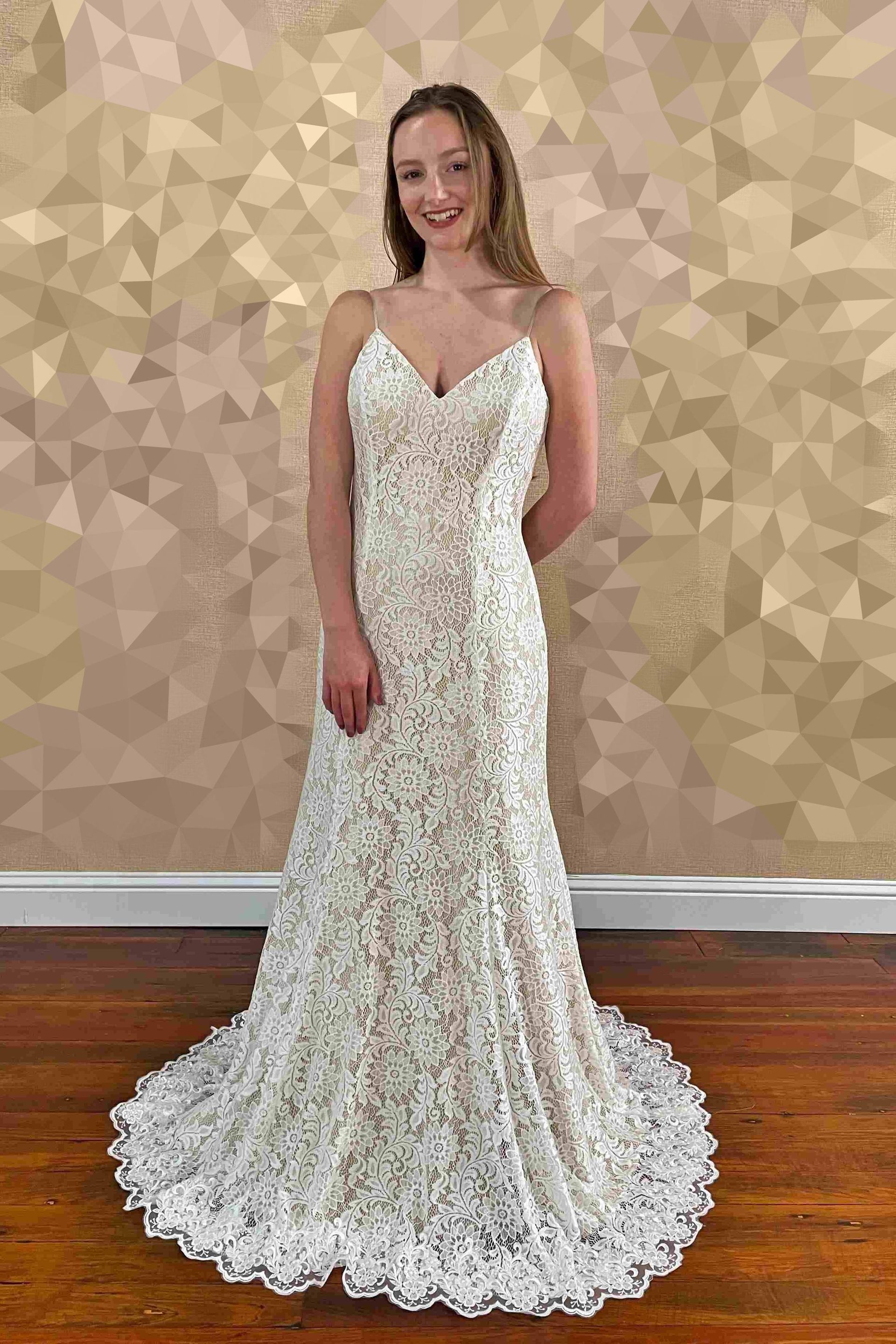 Woman in a white lace wedding dress smiles, standing indoors against a patterned wall.
