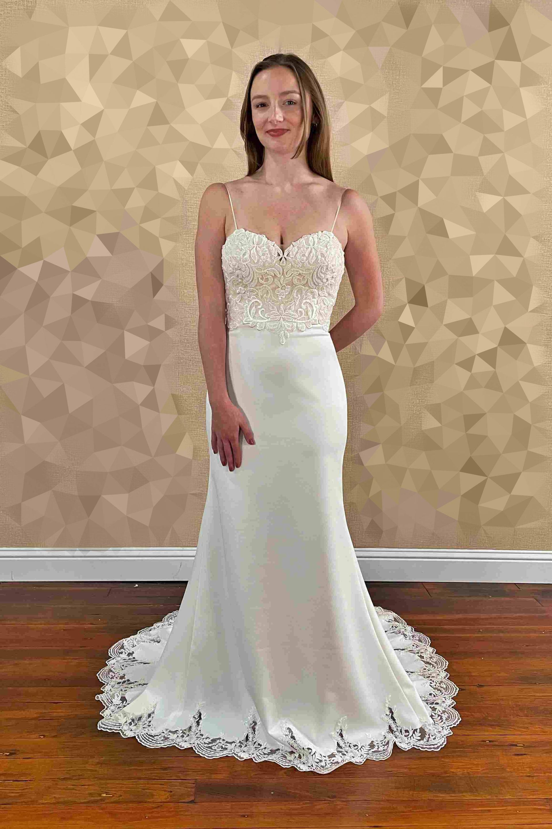 Woman in a white wedding dress with a decorated bodice and lace trim. Standing in front of a textured wall.