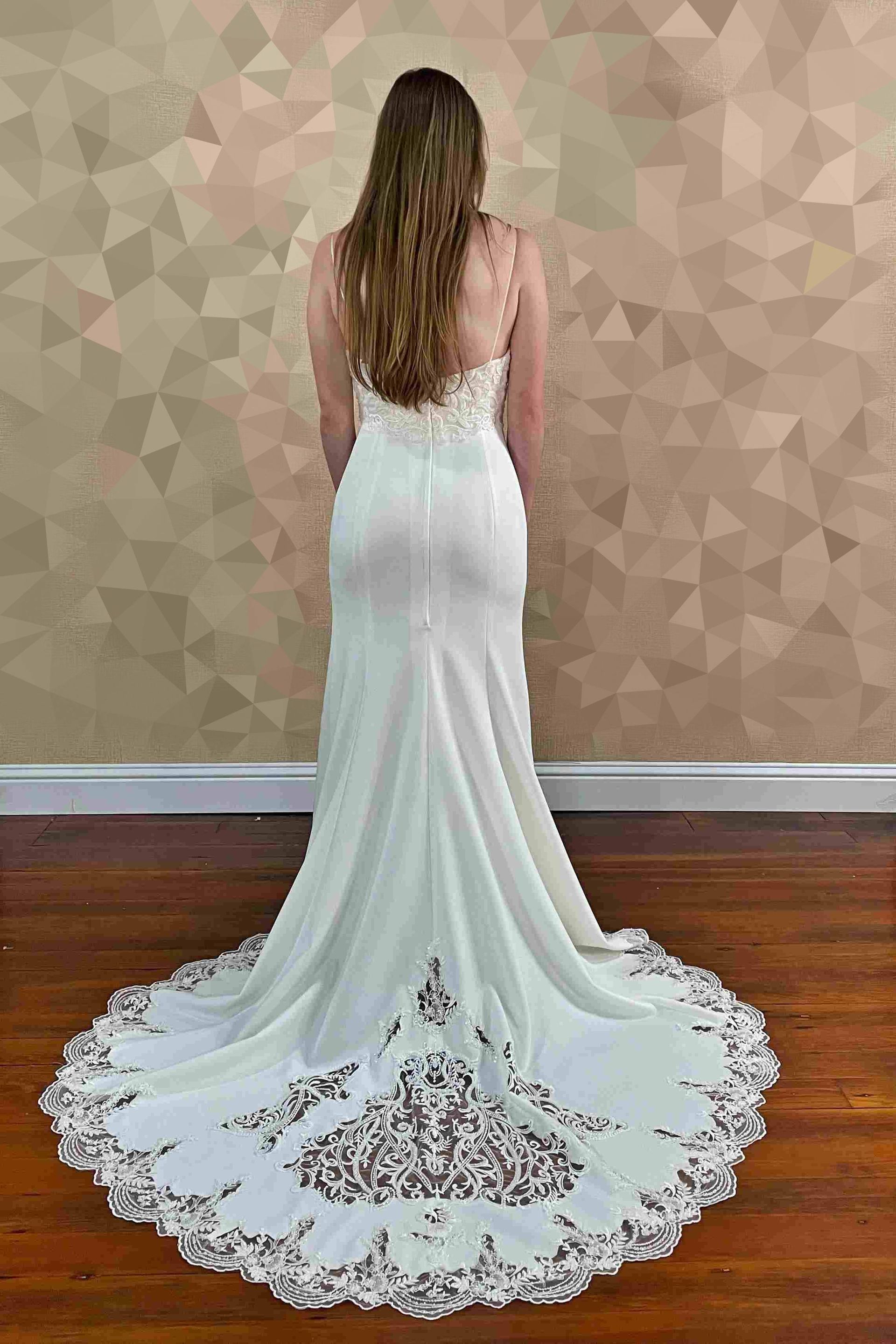 Woman in white wedding dress with long train, standing against patterned wall.