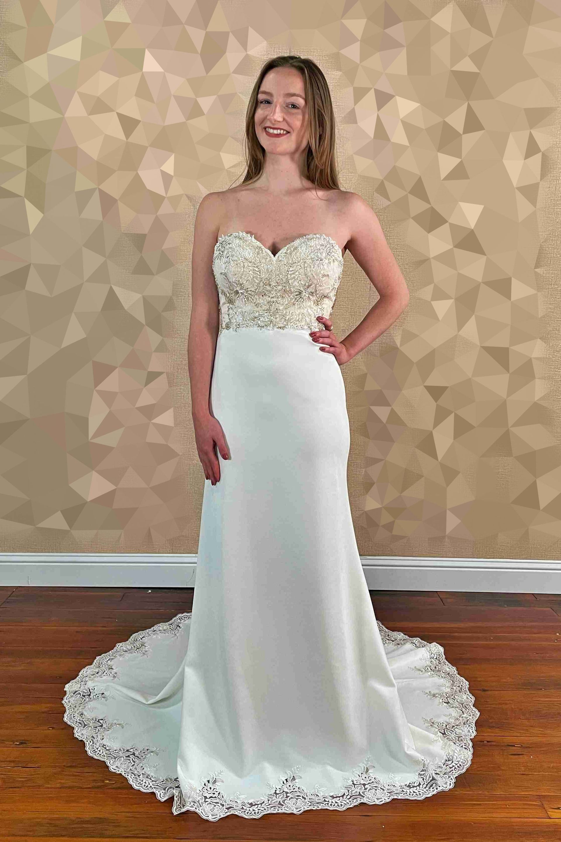 Woman in a strapless white wedding dress with beaded bodice, train, and hand on hip, smiling.