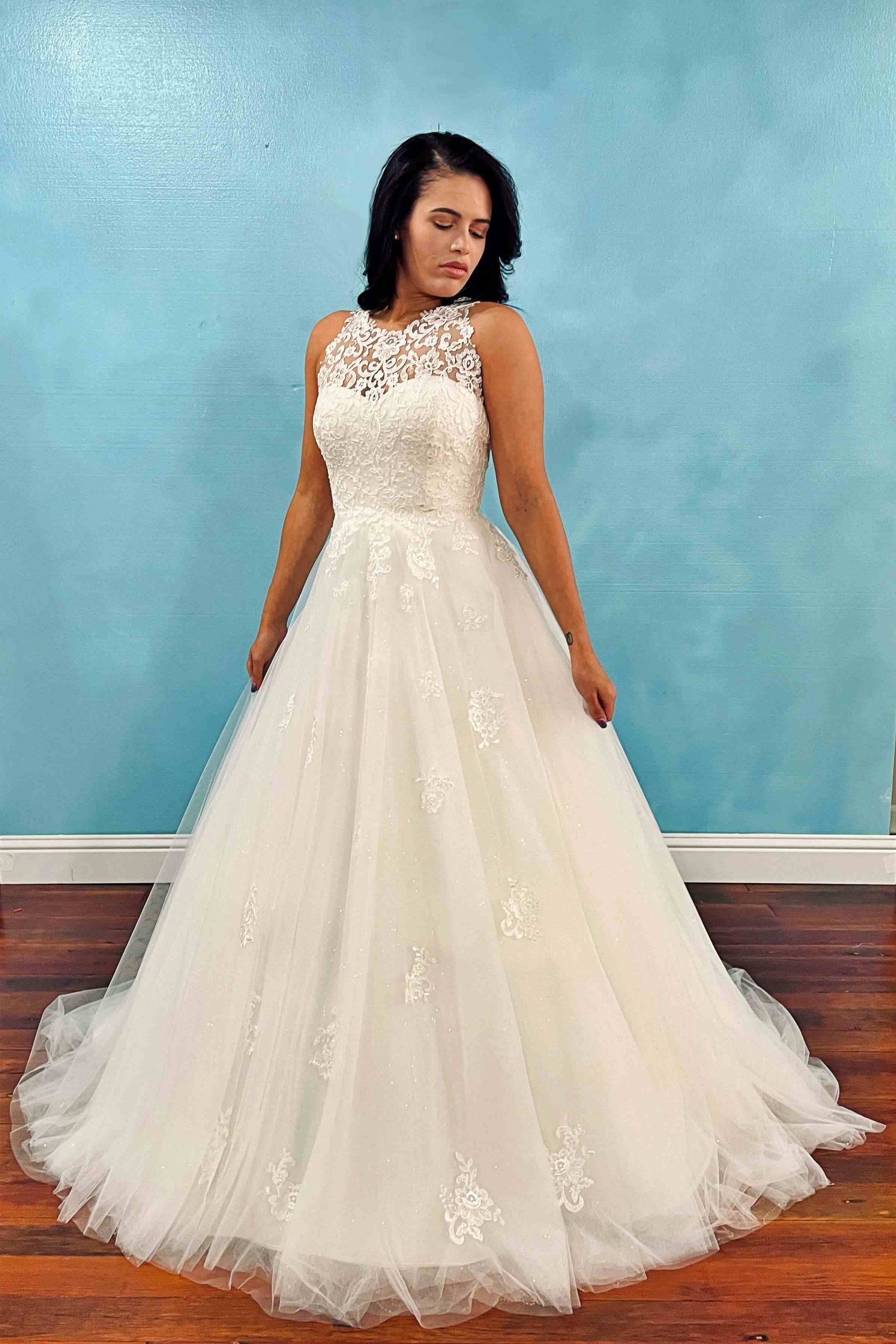 Woman in a white wedding gown with lace bodice and full skirt, standing in front of a blue wall.