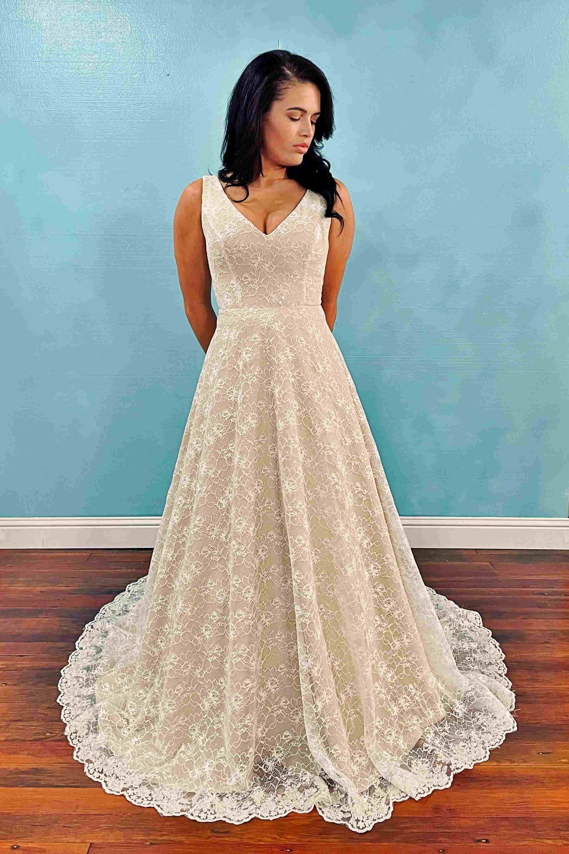 Woman in a white floral wedding dress stands, hands behind her back, in front of a blue wall.