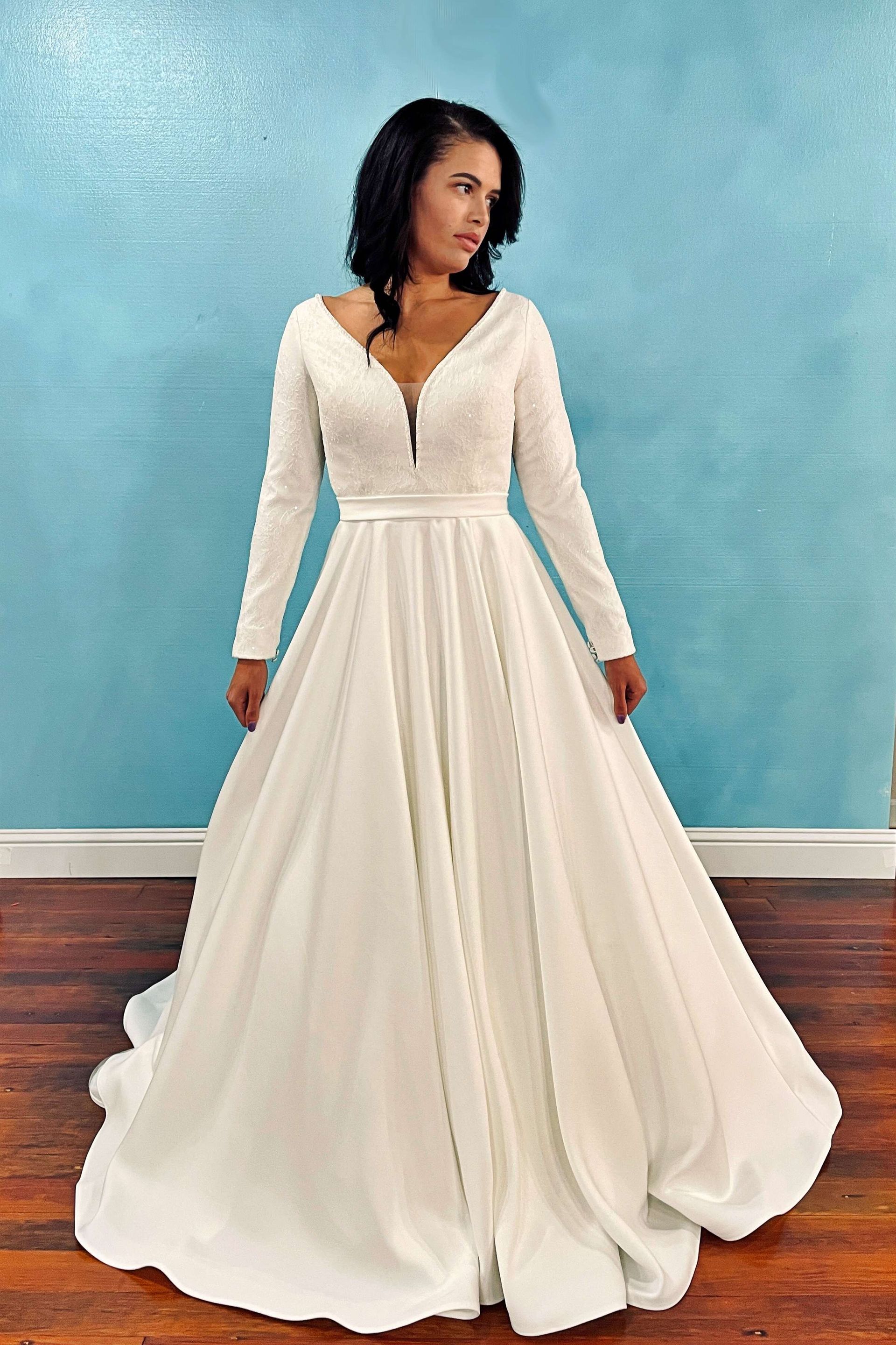 Woman in a white long-sleeved wedding dress with a low neckline, standing in front of a blue wall.