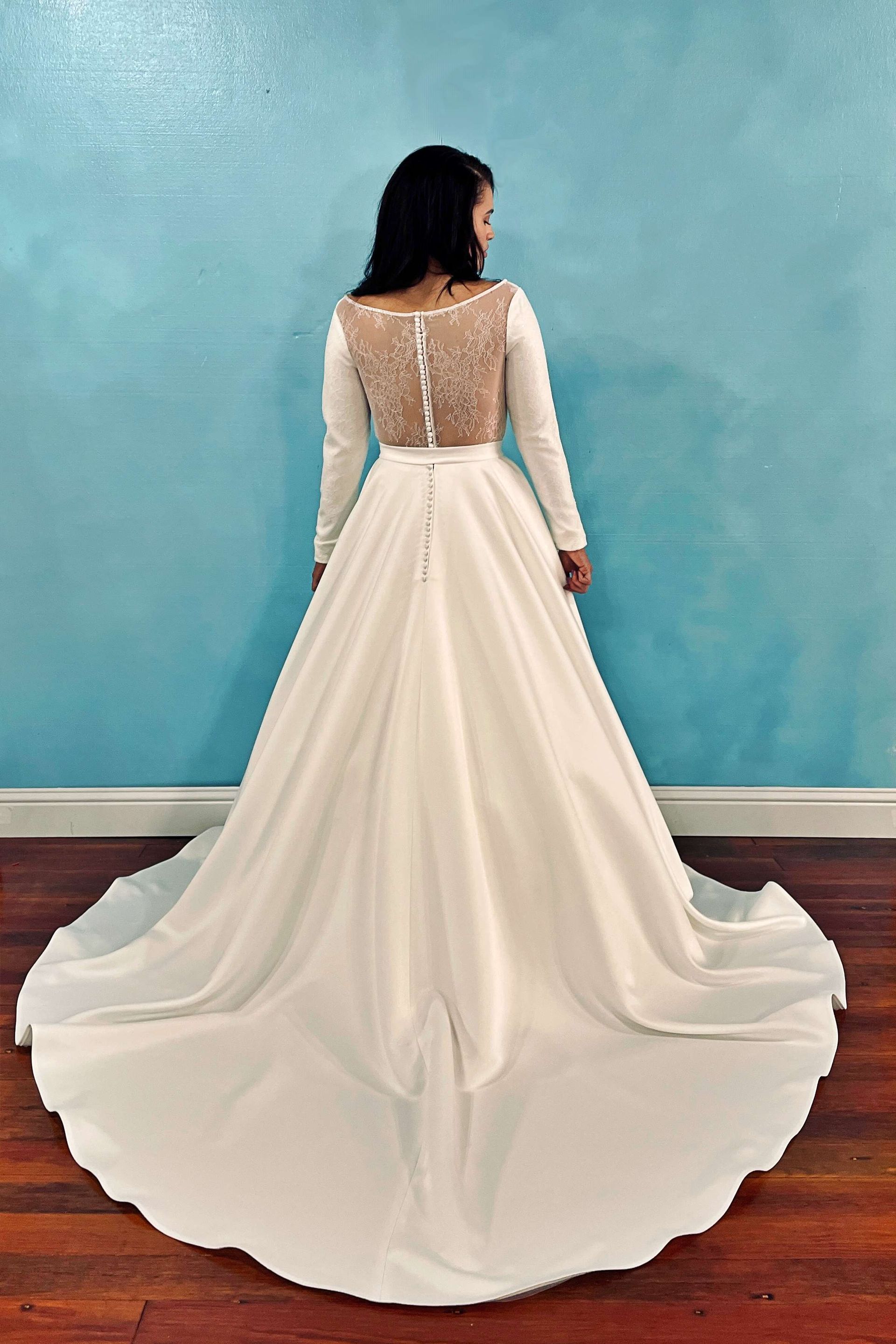 Woman in white wedding dress with long train, back view. Dress has long sleeves and sheer embellished back.
