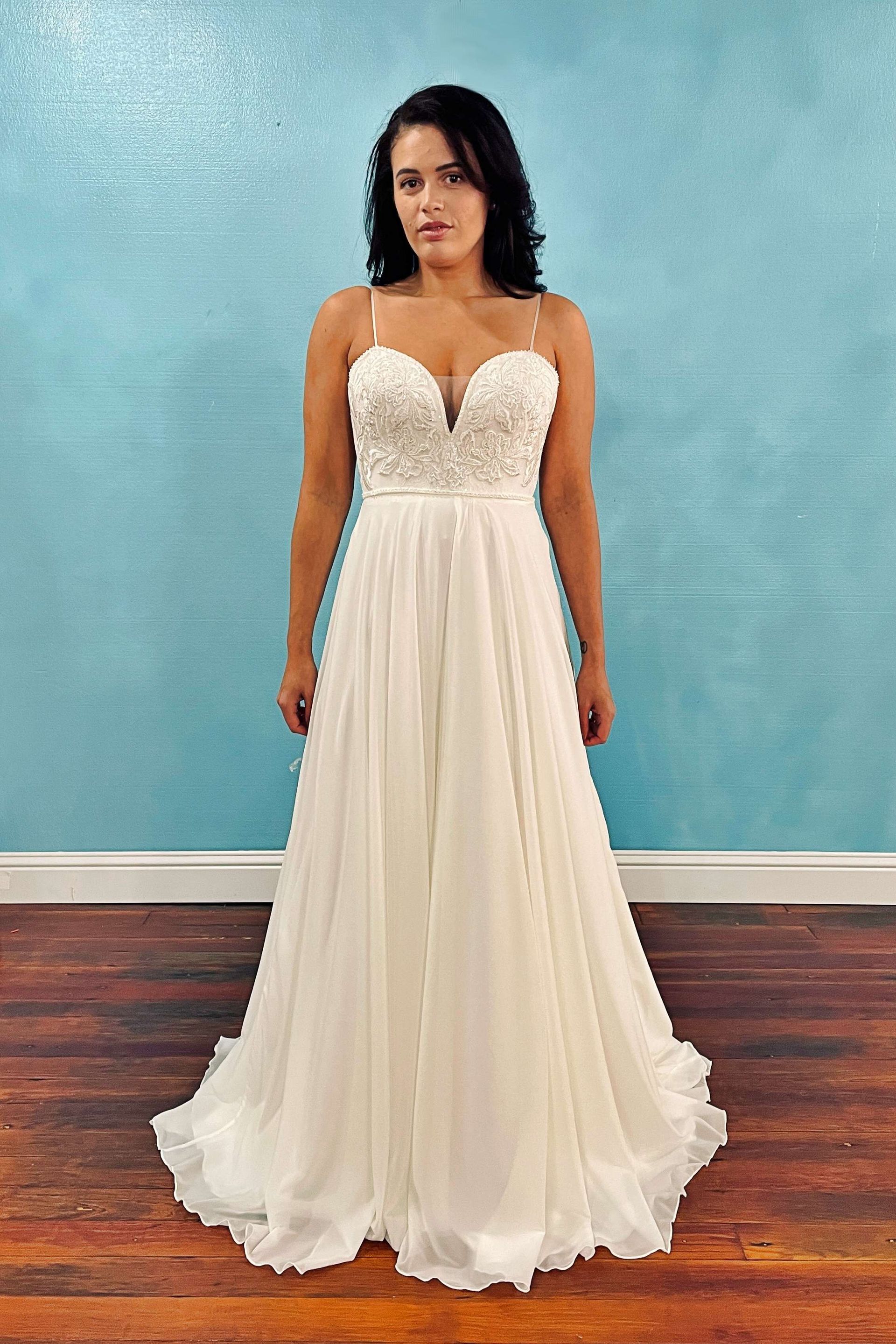 Woman in a white wedding dress with floral bodice and flowing skirt, standing in front of a blue wall.
