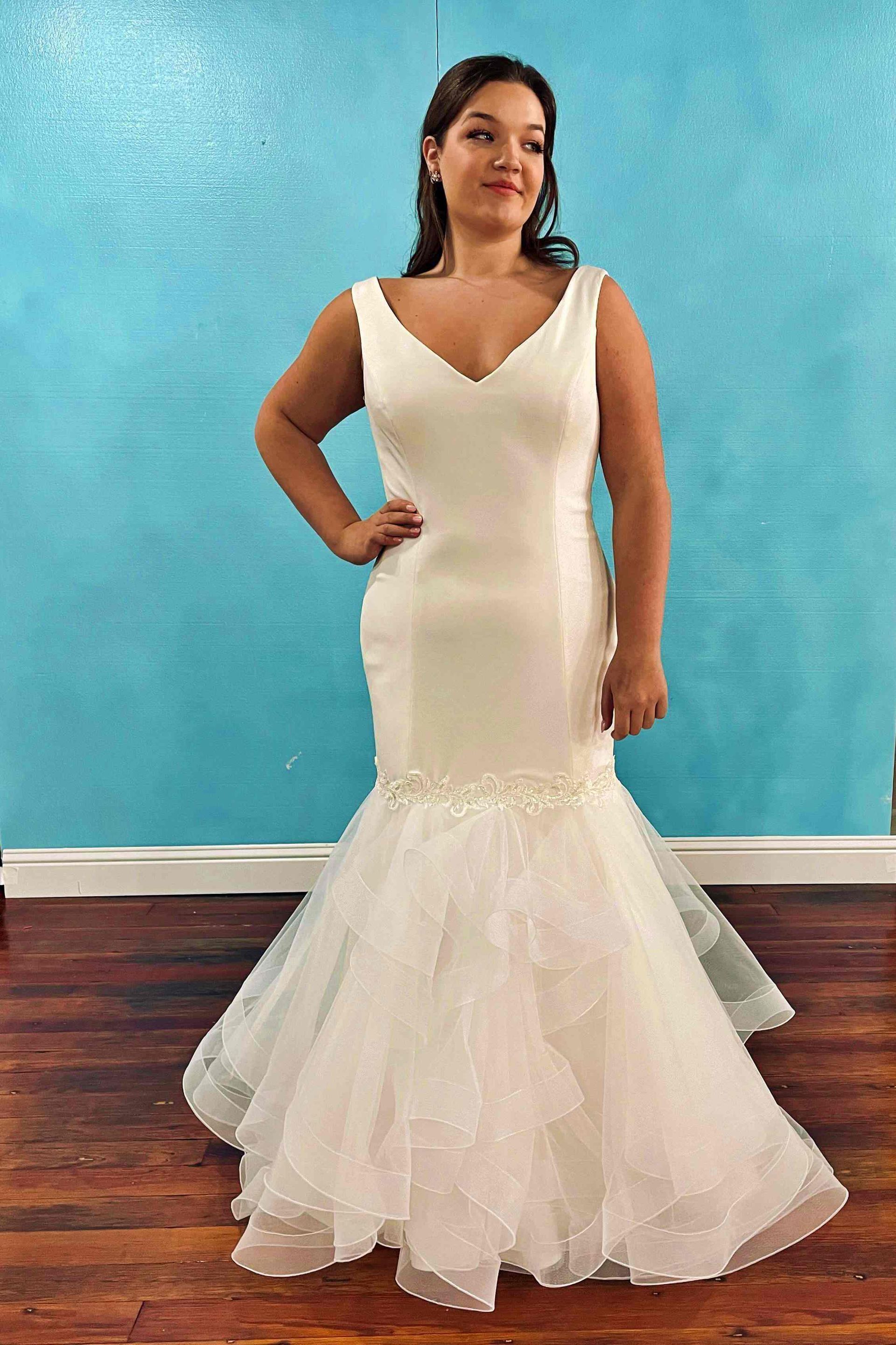 Woman in white wedding dress with V-neckline, mermaid skirt, hand on hip, in front of a blue wall.