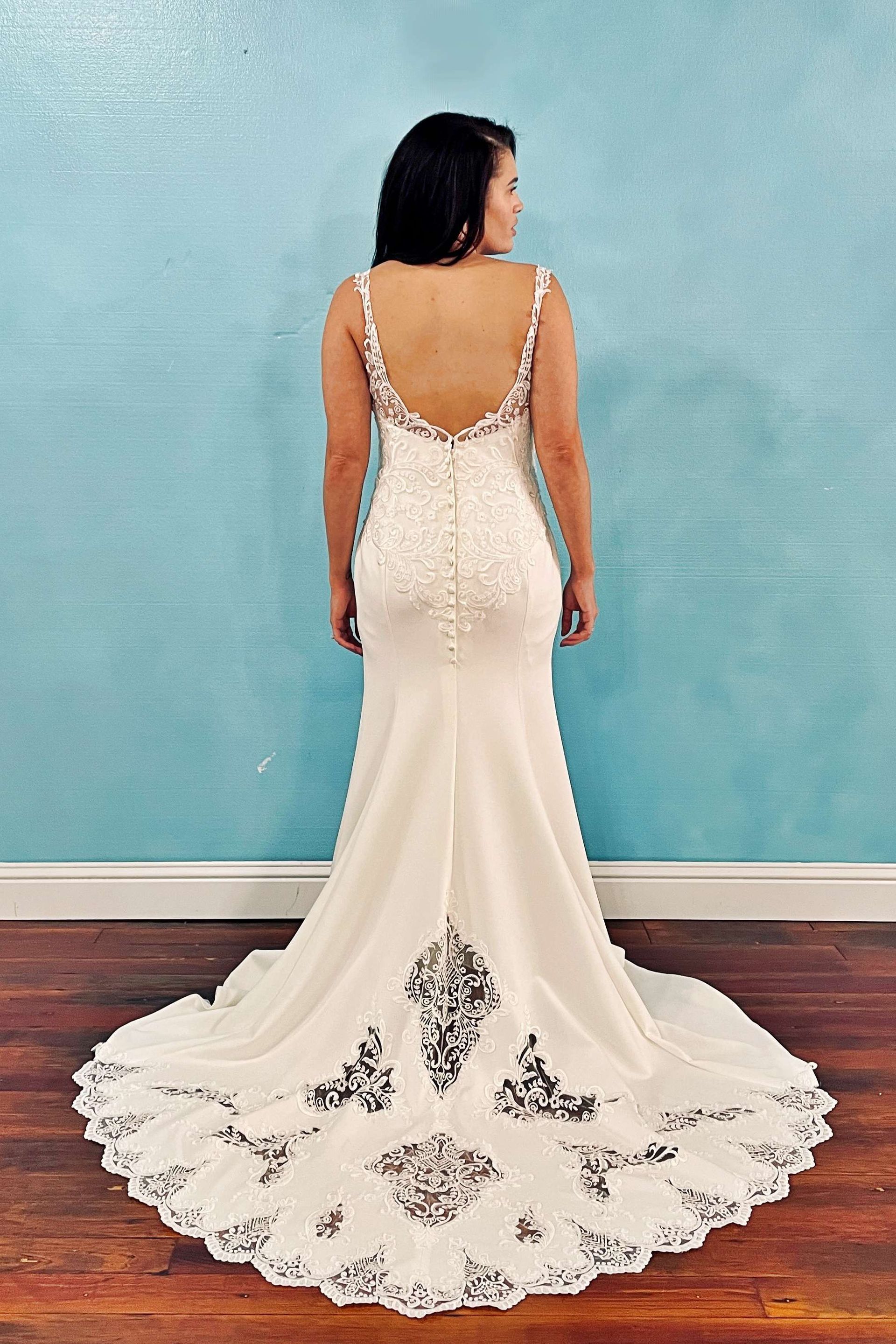Woman in a white wedding gown with a train, standing against a light blue wall. Dress has black lace details and a low back.